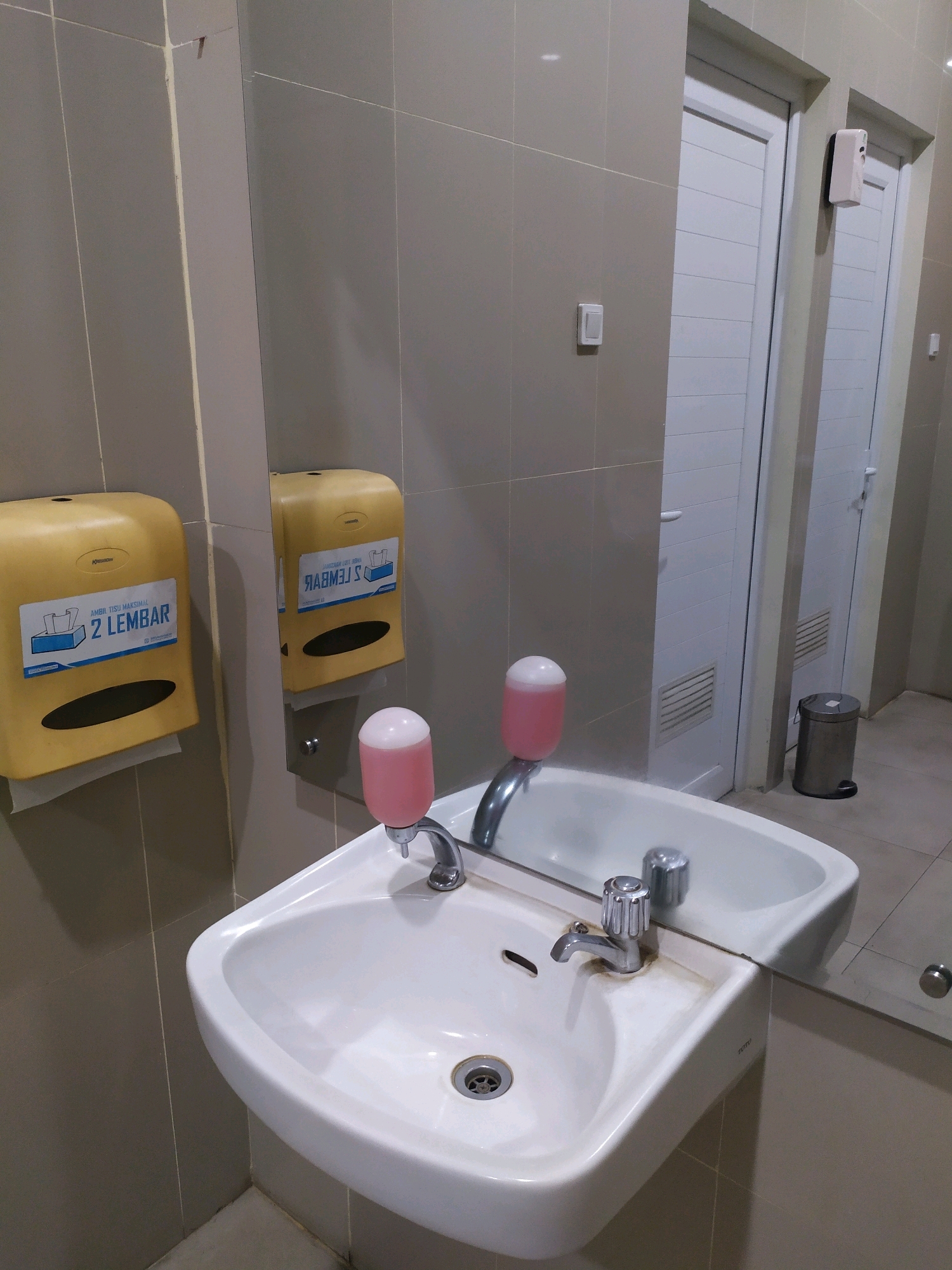 The image shows a clean, well-lit public restroom in Sleman, Indonesia. A white ceramic sink with a chrome faucet is positioned in the foreground, with a pink liquid soap dispenser mounted above it. To the left of the sink is a yellow tissue dispenser with Indonesian text that reads "AMBIL TISU MAKSIMAL 2 LEMBAR," indicating a maximum of two tissues can be taken. This dispenser is also reflected in a large mirror that covers a significant portion of the wall behind the sink. The walls are tiled with light gray tiles. In the background, there are two white doors, likely leading to toilet stalls or other rooms. A silver trash can is visible to the right of the doors. The floor is tiled with light brown tiles. The overall impression is of a functional and maintained restroom facility.