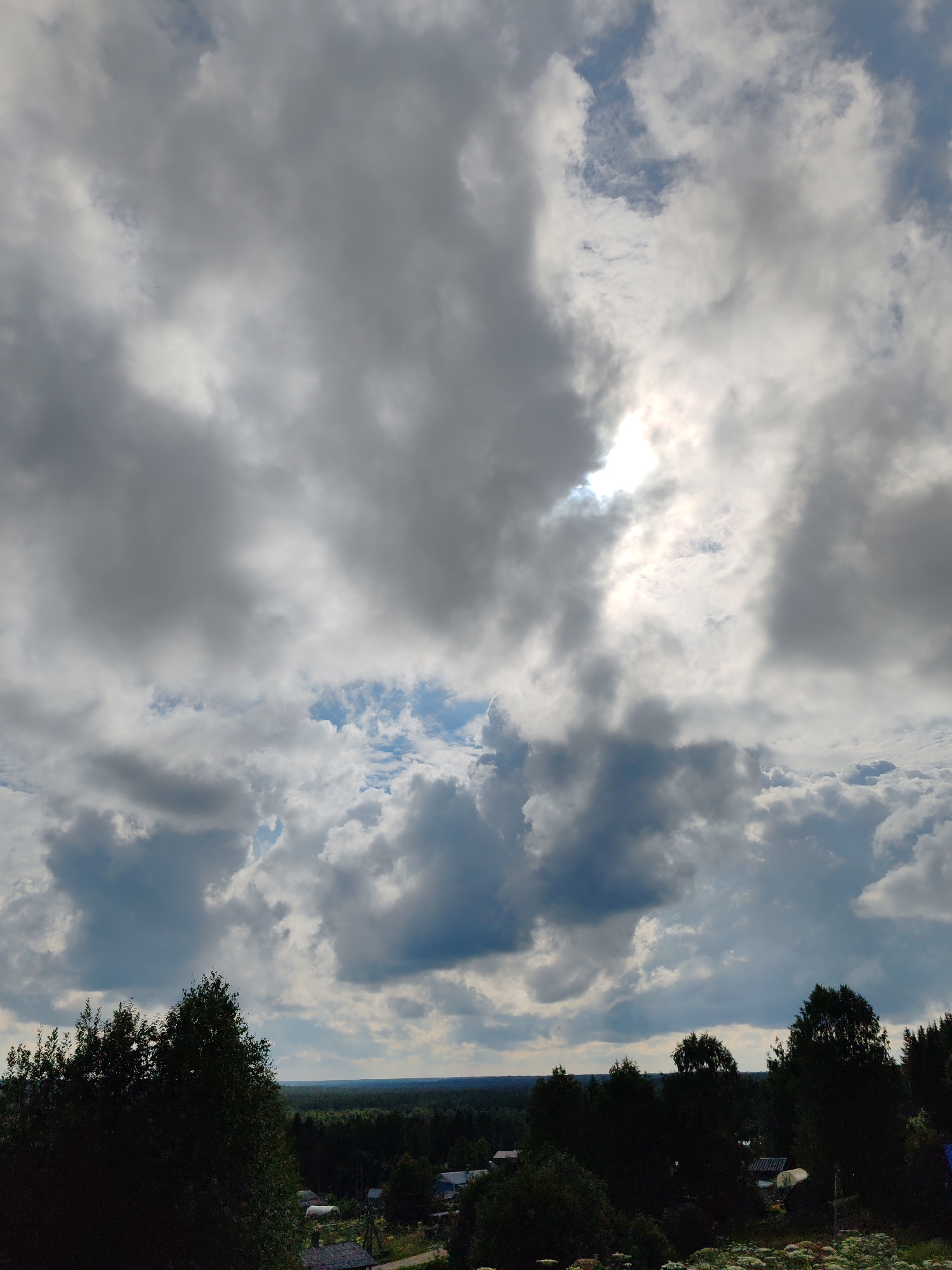The image depicts a cloudy sky over a rural landscape in Chukhlem, Russia. The sky is filled with large, gray and white clouds, with patches of blue visible between them. Sunlight is breaking through the clouds in the upper right portion of the image, creating a bright glow. Below the sky, a dense forest stretches across the horizon. In the foreground, a few trees are silhouetted against the sky, and some small buildings or houses are visible amidst the trees. The overall impression is one of a natural, somewhat overcast environment. There are no people visible in the image.