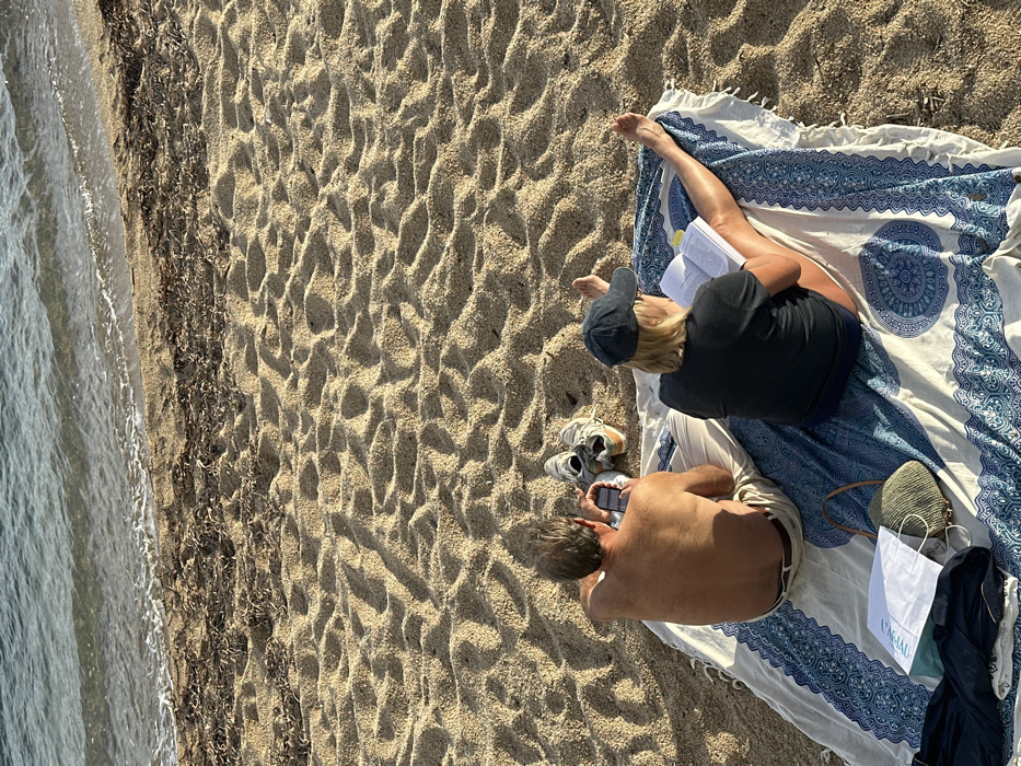 On a sandy beach in Palafrugell, Spain, two individuals are seated on a patterned blue and white blanket, observed from behind. The male, positioned on the left, is shirtless and wears light-colored trousers, holding and interacting with a smartphone. Behind him on the sand is a pair of light-colored sneakers. The female, on the right, wears a dark t-shirt and a dark baseball cap, reading an open book. Her bare legs are extended on the blanket. Adjacent to the male's left side on the blanket are a greenish-beige woven bag, a white paper bag, and a dark piece of fabric. The sandy environment features ripples and depressions, with a line of dark organic debris along the waterline, beyond which calm, light blue-green ocean water is visible under a warm, low-angle light.