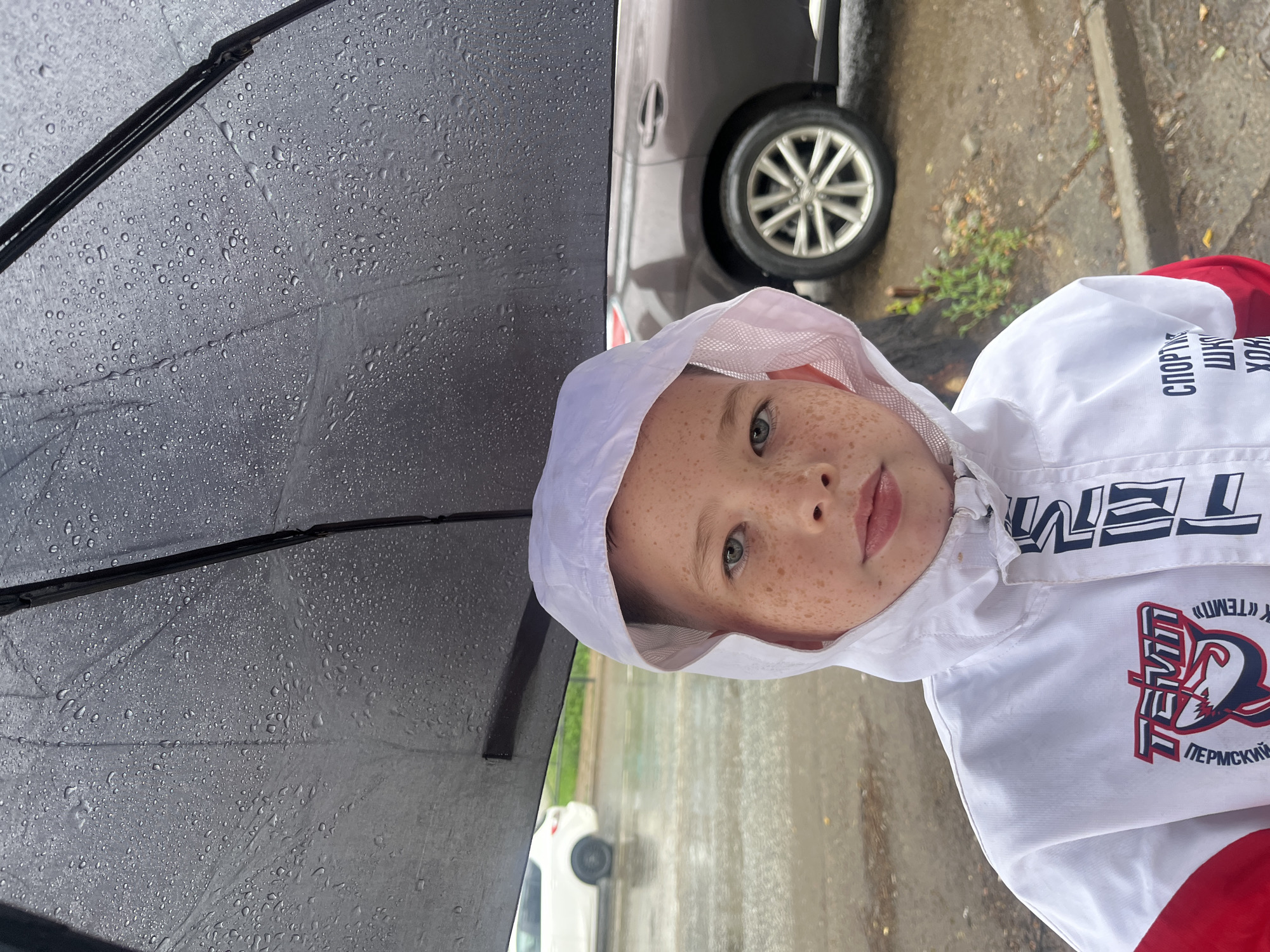 A young boy with freckles stands under a dark umbrella in the rain. He is wearing a white hooded jacket with red accents and sports logos. In the background, two cars are parked on a wet, paved surface, suggesting an outdoor urban environment. The scene appears to be in the city of Chaikovsky, Russia, as indicated by the Cyrillic text on the boy's jacket, which includes "ПЕРМСКИЙ" (Permsky) and potentially references to a sports school related to hockey. The wet asphalt and raindrops on the umbrella confirm it is raining.