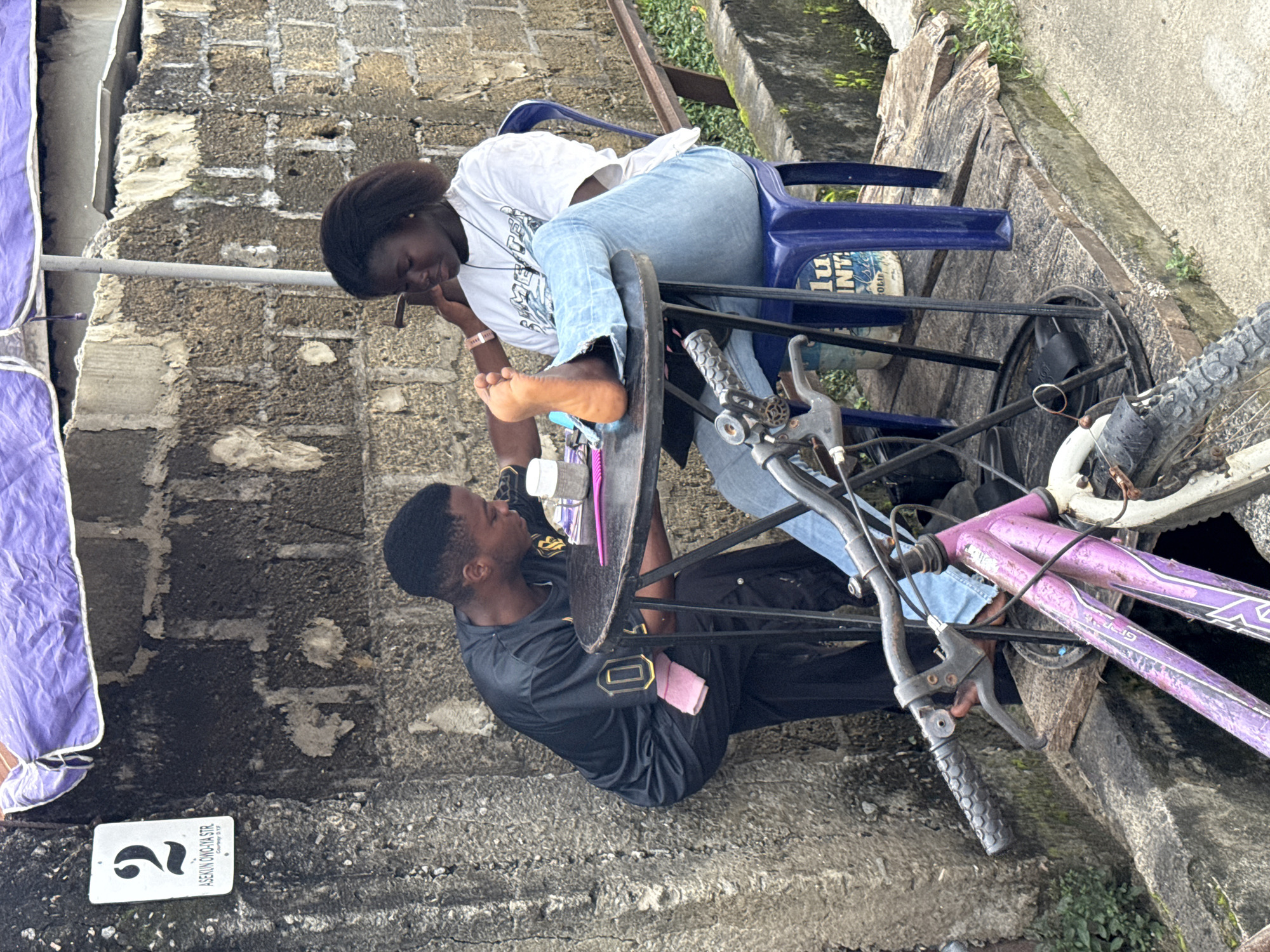 In Ado, Nigeria, a young man in a black t-shirt and black pants is bent over a small, round, black metal table, attending to the foot of a young woman seated in a blue plastic chair. The woman is wearing a white t-shirt and light blue jeans, with her right leg extended and her foot resting on the table. The man is holding a tool and appears to be working on her toenails. A pink cloth is tucked into his waistband. On the table are several small items, including a white cylindrical container and a pink utensil. The scene is set against a textured, grey brick wall, partially covered by a purple tarp. A white sign with the number "2" and "ASEKUN OWO-IYA STR" is visible on the wall to the left. In the foreground, a portion of a purple bicycle is visible, with its handlebars and front wheel in view. The ground is a mix of concrete and wooden planks. Greenery is present in the background.