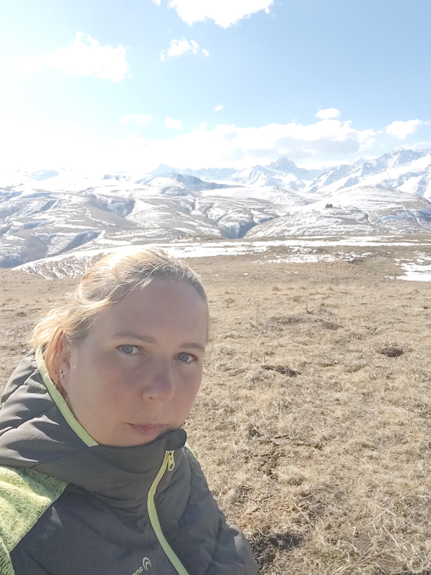 A blonde woman is taking a selfie in an outdoor setting with a mountain range in the background. The mountains are partially covered in snow and the sky is light blue with scattered clouds. The foreground consists of dry, brown grass and patchy snow. The woman is wearing a dark grey jacket with a lime green accent on the collar and sleeves. The brand "Karuno" is visible on the jacket. The location is likely near Tyrnyauz, Russia, given the mountainous terrain and snowy conditions.