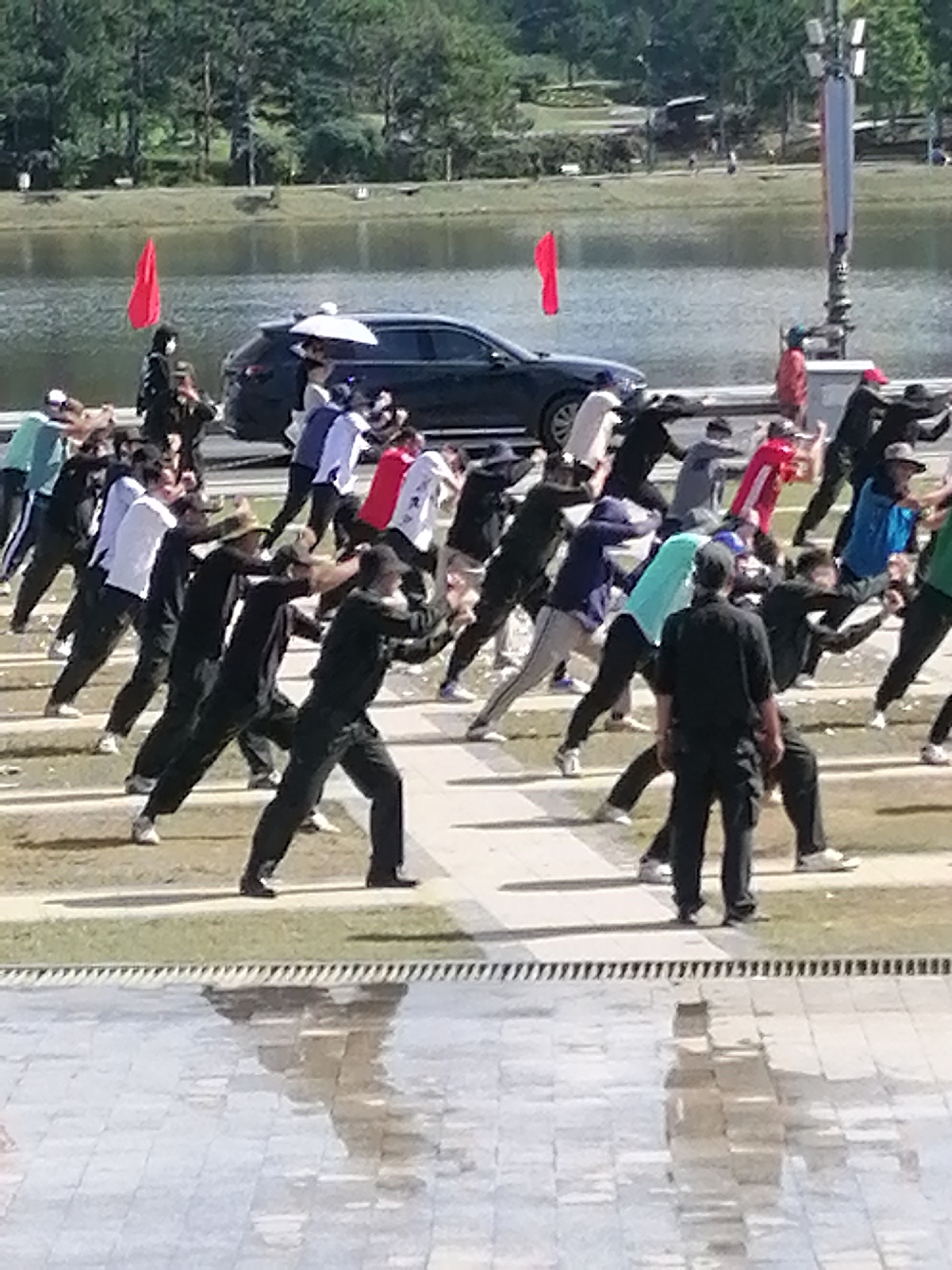 The image shows a group of people engaged in what appears to be a tai chi or similar slow-movement exercise session outdoors. The subjects are predominantly men, dressed in casual athletic wear, mostly dark-colored pants and a variety of shirts in shades of blue, white, red, and green. They are positioned on a paved area, possibly a plaza or promenade, with a body of water and a tree-lined background suggesting a park or lakeside setting.
In the mid-ground, a dark-colored car is parked, and a person is holding a white umbrella over another individual. Two red flags are visible on poles, one near the car and another further back on the left, possibly indicating a specific event or location. The overall atmosphere seems calm and focused, with the participants exhibiting synchronized movements.
The lighting suggests it is daytime, likely late morning or afternoon, given the shadows cast on the ground. The weather appears to be clear and sunny, contributing to the outdoor activity. The presence of water and lush greenery points to a scenic location, consistent with the provided context of Da Lat, Vietnam. Reflections are visible on the wet paved surface in the foreground, adding a visual element to the scene. No visible text is present in the image.