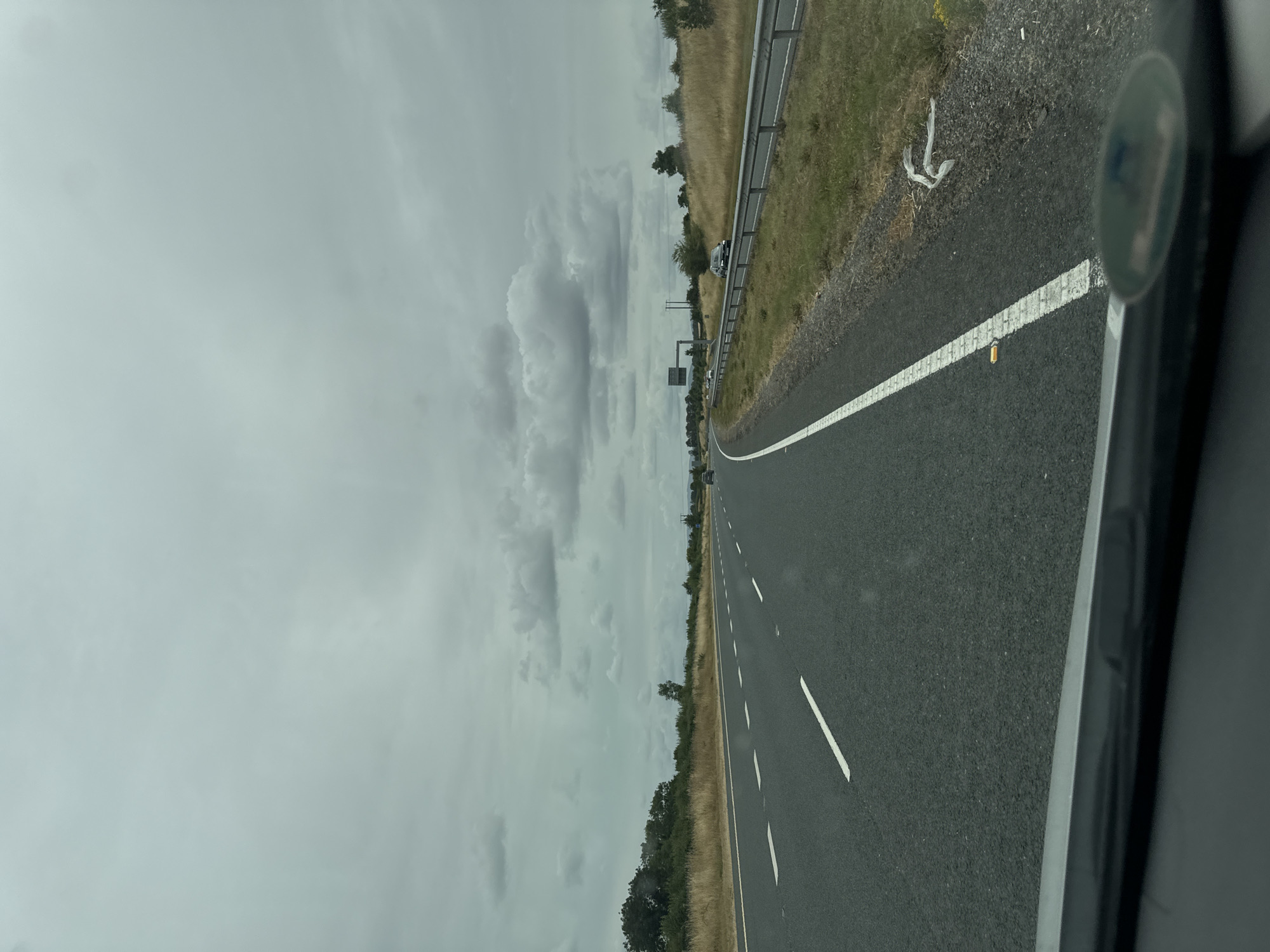 The image captures a view from a vehicle traveling on a highway, likely the M8 motorway, in Knockraheen, Ireland. The road is asphalt with clear white lane markings. The sky is overcast with dense, grey clouds, suggesting a potentially gloomy day. On the right side of the road, a metal guardrail separates the highway from a grassy verge dotted with dry, yellow grass and some scrubby bushes. Further in the distance, trees and more vegetation line the horizon. A few cars are visible on the highway, one in the distance on the right lane and another on the left lane ahead. Near the edge of the road on the right verge, a white plastic bag is visible, along with some scattered debris. The overall impression is that of a rural highway scene under a cloudy sky.