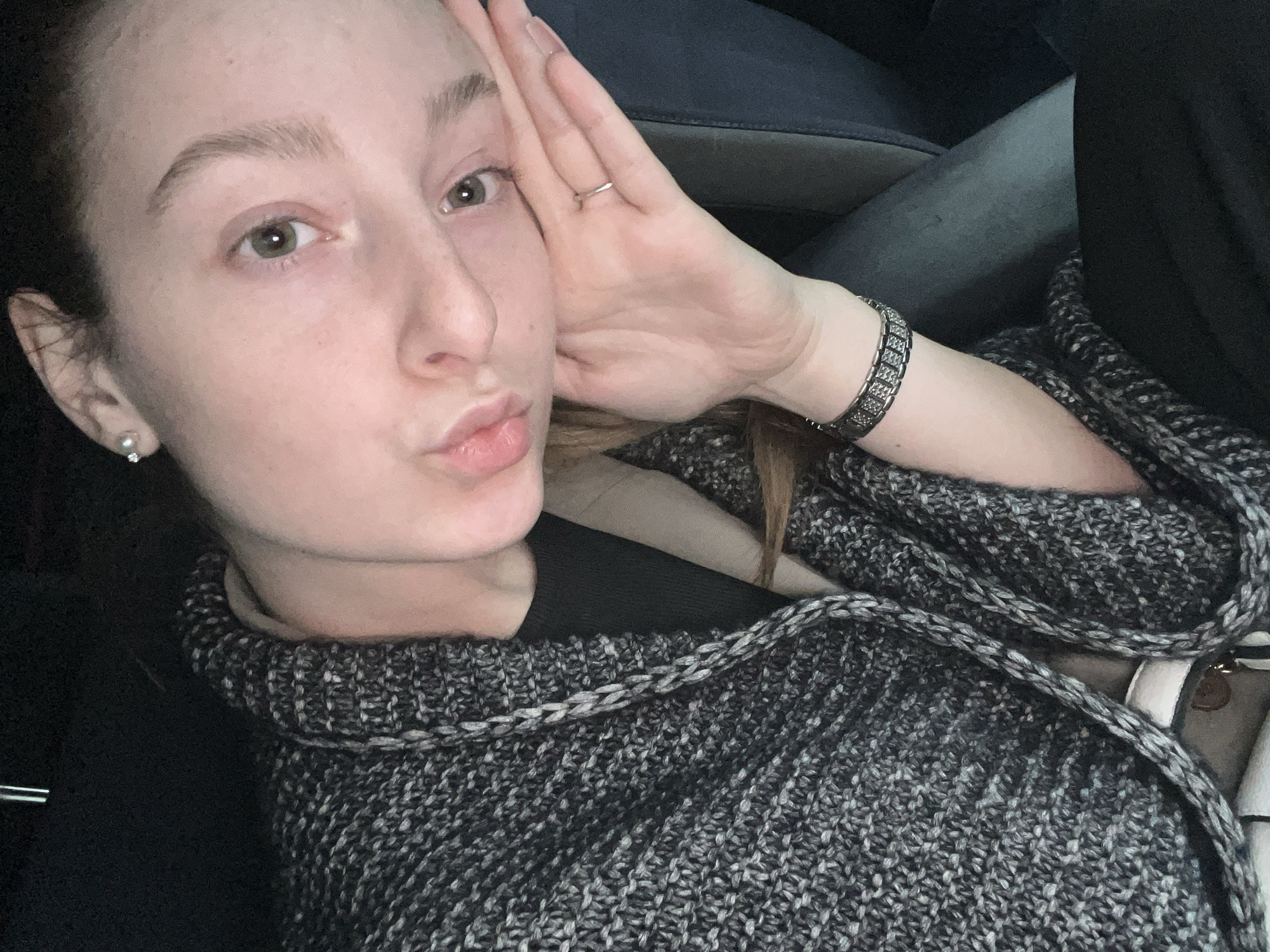 A selfie taken indoors shows a fair-skinned woman with light brown hair pulled back. She is wearing a gray and black knit sweater or shawl. Her left hand is resting against her forehead, and she is pouting her lips slightly. She has a silver ring on her finger and a decorative silver bracelet on her wrist. A small, pearl-like earring is visible in her left ear. The woman is sitting in what appears to be a car, with a gray car seat and black interior elements visible. The location of the photo is not possible to determine from the image provided, and Varna, Bulgaria cannot be confirmed.