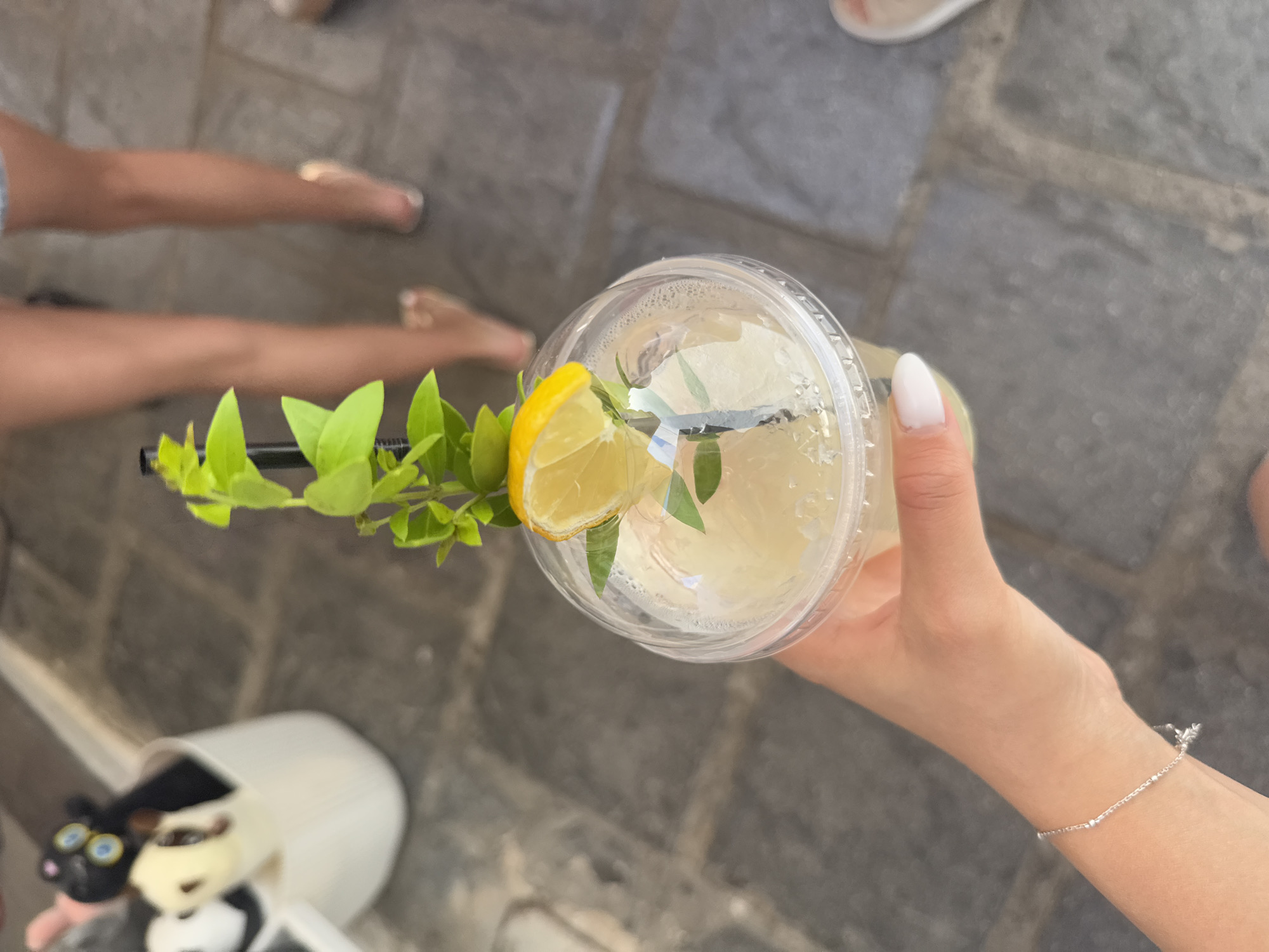 In Lindos, Greece, a person's hand, adorned with a delicate silver bracelet and a white-tipped manicure, holds a clear plastic cup filled with a pale yellow beverage. The drink is garnished with a slice of lemon, sprigs of fresh green leaves, and a black straw. The cup is sealed with a clear plastic lid, and condensation has formed on its surface. The background is a textured gray stone pathway, with blurred figures of people in the distance, suggesting an outdoor setting. To the left, a white container holds several plush toys, including a black one with large blue eyes and a cream-colored one. The scene captures a moment of refreshment, likely in a tourist area.
