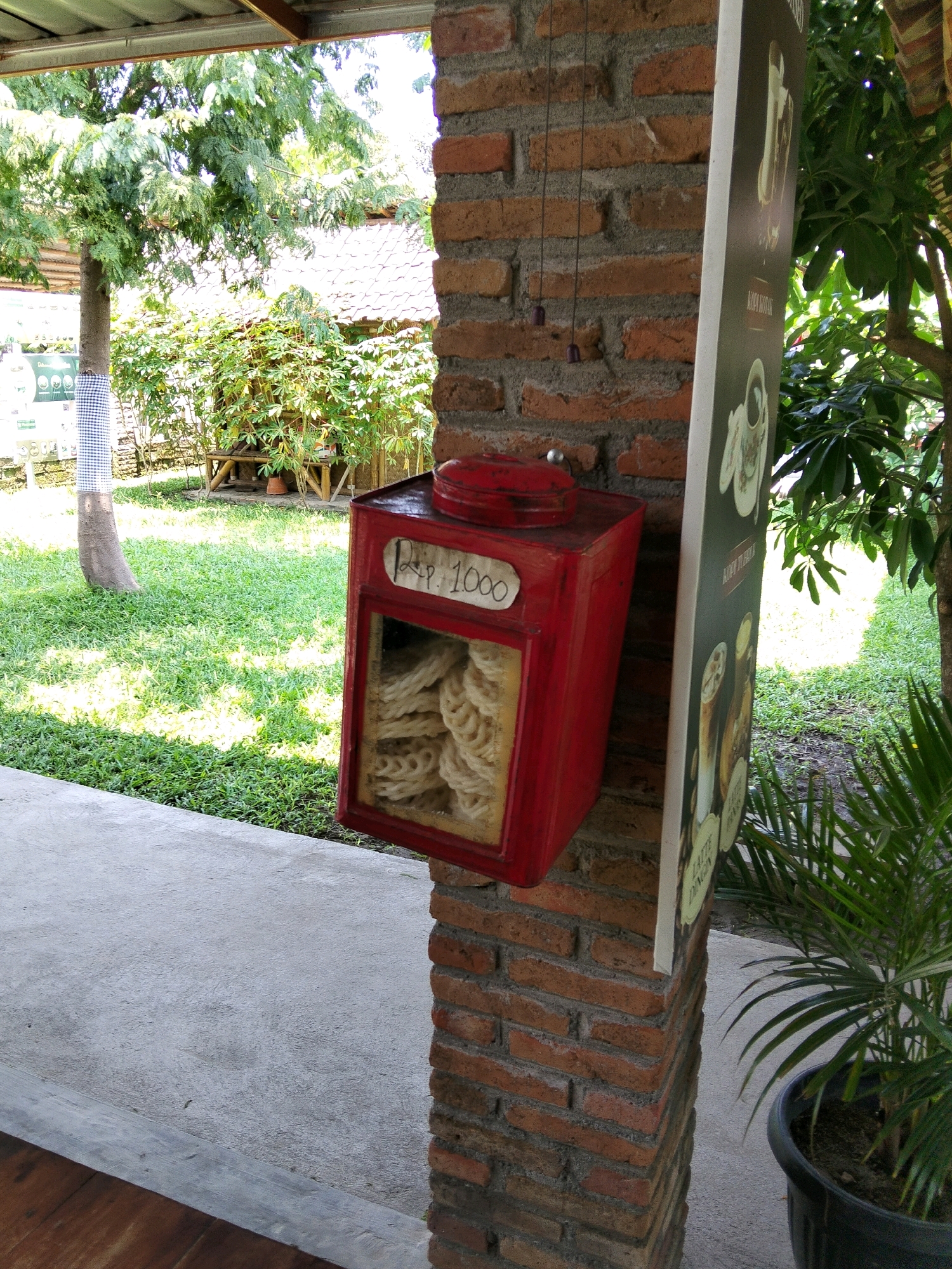 A red tin box filled with white crackers is attached to a brick pillar, with a sign that reads "Rp. 1000" on its front. The pillar is part of a building's exterior in Gurah, Indonesia, and is surrounded by lush green grass and trees. To the right of the pillar, a potted palm tree stands near a paved pathway. In the background, a traditional Indonesian building with a tiled roof can be seen amidst dense foliage. The scene appears to be an outdoor dining or recreational area.