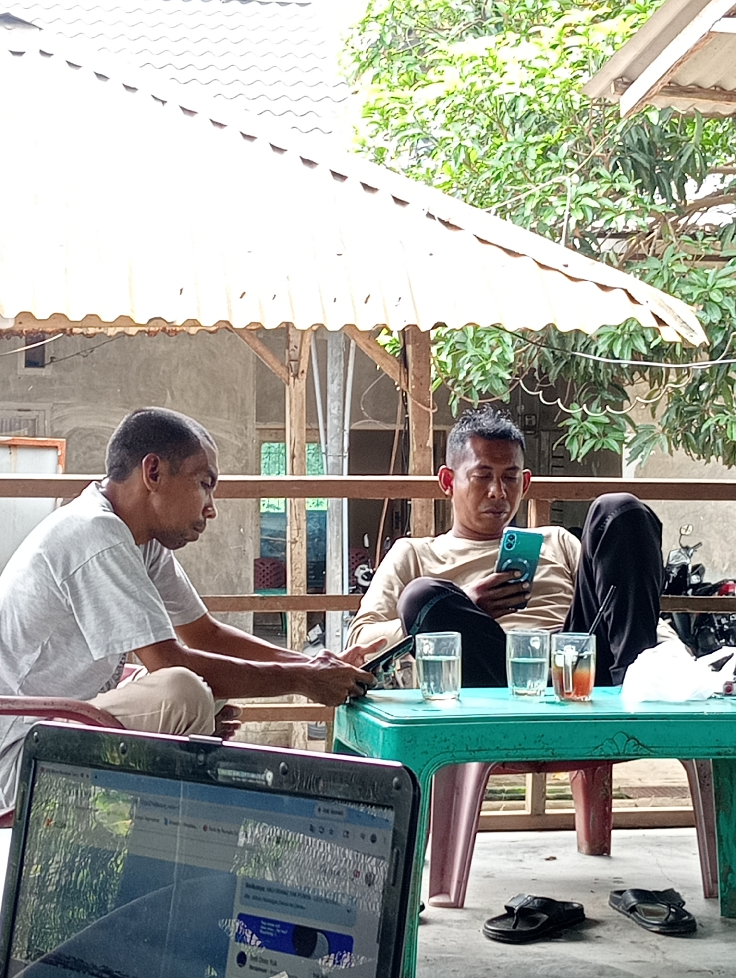 Two men are seated at a green plastic table on a porch or patio. One man, wearing a white t-shirt and tan pants, sits to the left and appears to be focused on something partially obscured by a laptop in the foreground. The other man, wearing a tan long-sleeved shirt and black pants, is leaning back in his chair, looking at a teal-colored smartphone. Three glasses are on the table, one filled with a dark-colored liquid. A pair of black flip-flops lies on the floor beneath the table. The background includes a white roof overhang, a building exterior, trees, and a motorbike. The location appears to be Lhokseumawe, Indonesia.
