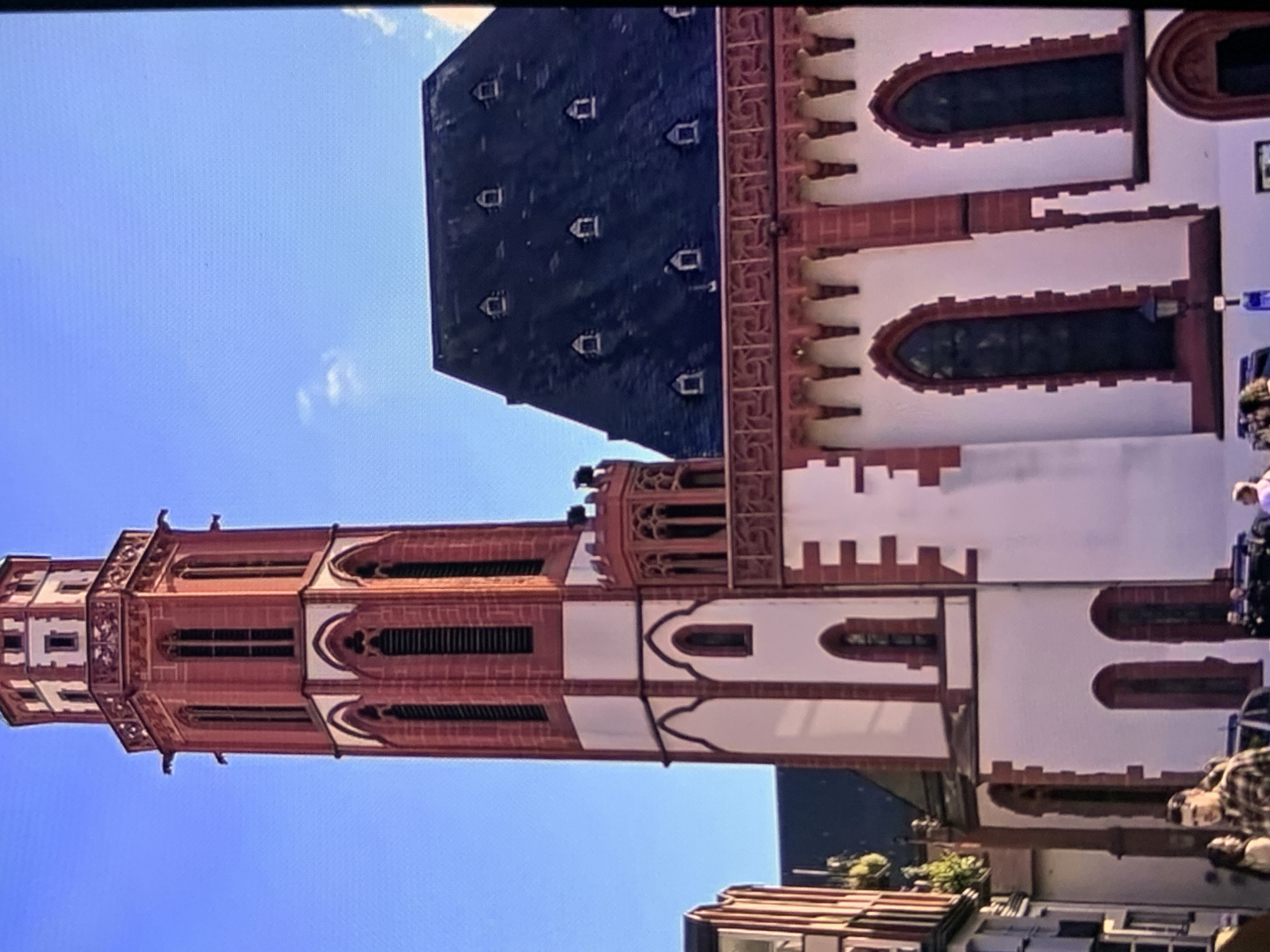 This media file is a photograph of a tall, red-brick tower with white accents, likely part of a church or historical building. The tower has multiple levels with arched windows and decorative stonework. A dark, steeply pitched roof with small dormer windows is visible to the right of the tower, against a clear blue sky. The image is oriented vertically, with the tower appearing to stretch upwards from the bottom of the frame. The lighting suggests it is daytime, and the sky indicates fair weather. Some people are visible at the bottom right of the frame, appearing to be tourists or visitors. The overall scene depicts an architectural landmark in a city setting. No visible text is present in the image.