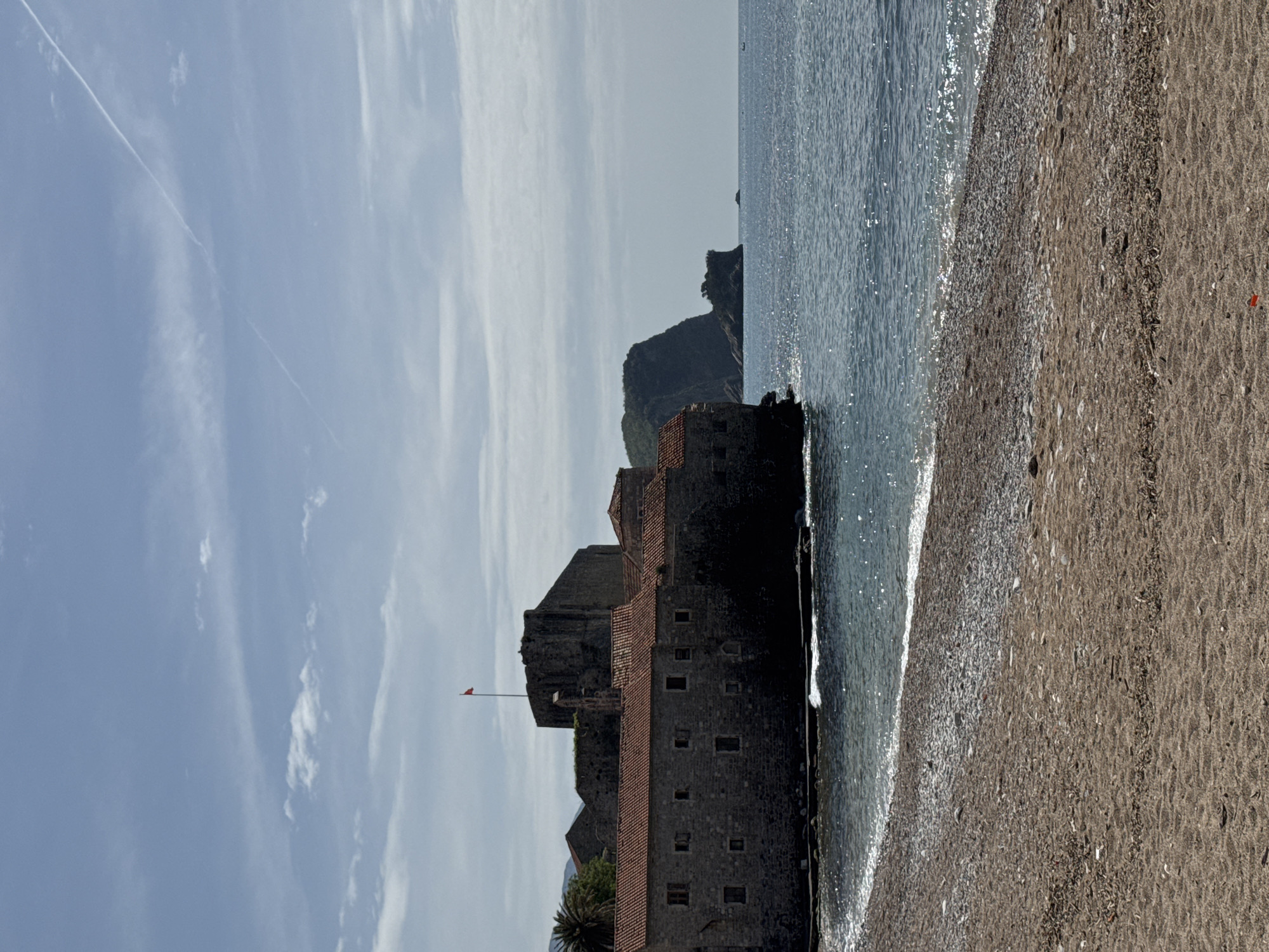 The image depicts a coastal scene in Budva, Montenegro, focusing on a stone building near the shoreline, likely part of the old town fortifications. The building is multi-storied with a red-tiled roof and small windows. A flag flies atop one of the towers.
The structure is situated at the edge of a beach, where waves are gently breaking. The beach itself appears sandy with small pebbles.
In the background, a rocky promontory juts out into the sea. The sky is pale blue with wispy clouds and contrails from aircraft.