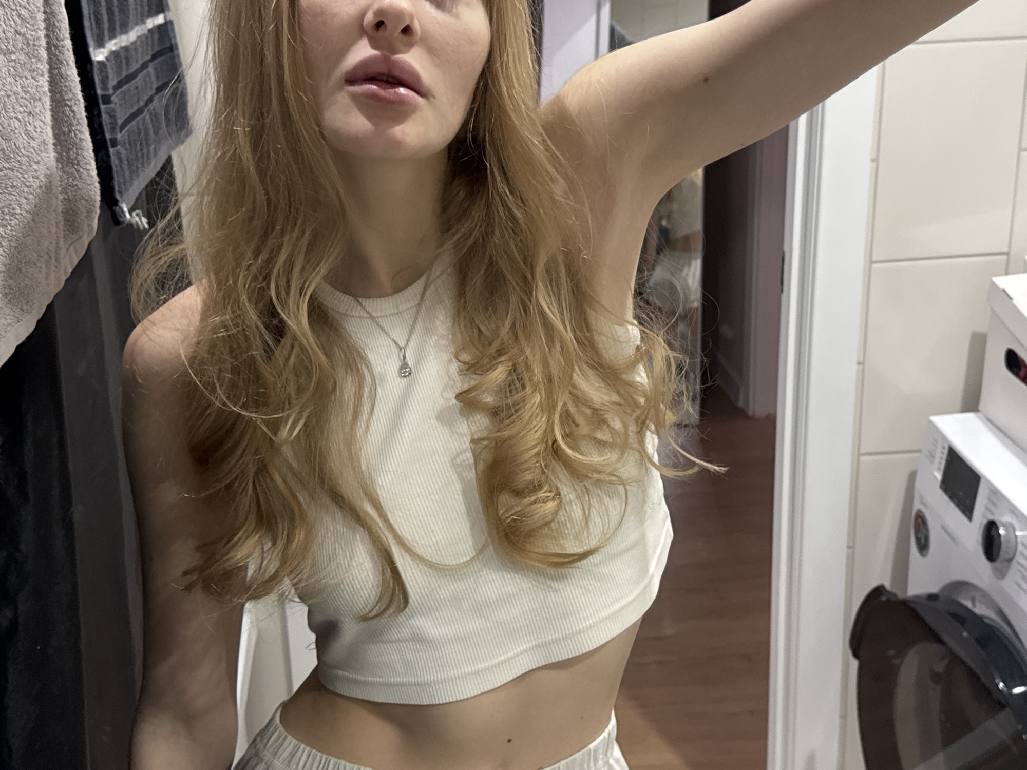 A blonde woman wearing a white tank top and light-colored bottoms is taking a selfie in a bathroom. A silver necklace is visible. The bathroom contains a washing machine and is possibly located in a home or apartment. It is impossible to confirm the location is in Perm, Russia from the image.