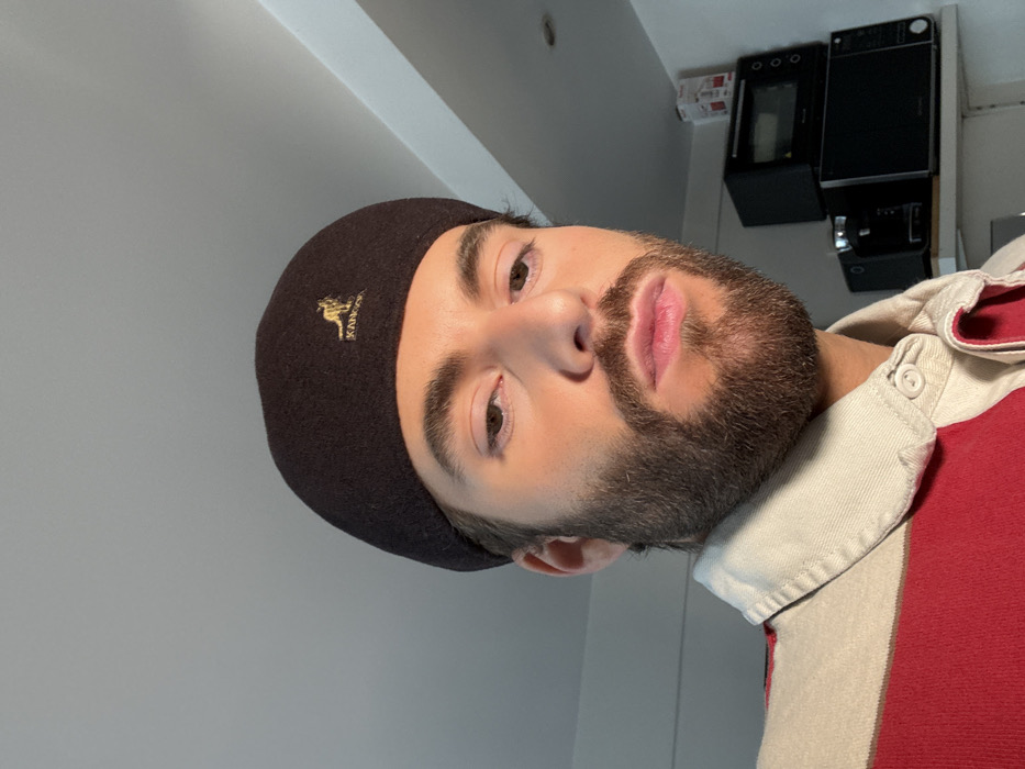 The image displays a close-up portrait of a Caucasian male, captured from the chest up against an indoor background. He has light brown eyes, dark brown hair visible at the temples, and a trimmed dark brown beard and mustache. The male wears a dark brown Kangol beret, featuring a gold embroidered kangaroo logo on the front-left side. His attire also includes a collared polo-style shirt with a cream-colored collar and broad horizontal stripes of cream and red. His lips are slightly parted, and his gaze is directed forward, towards the camera.
The background consists of a plain white wall or ceiling area. To the right of the subject, a white shelf is visible, supporting two stacked black kitchen appliances: a microwave oven on the bottom and a smaller toaster oven above it. A small white box with red markings is partially visible above the upper appliance. The lighting appears to originate from the left side of the frame. The indoor setting lacks specific identifying landmarks or architectural features to determine a precise location within Paris, France.