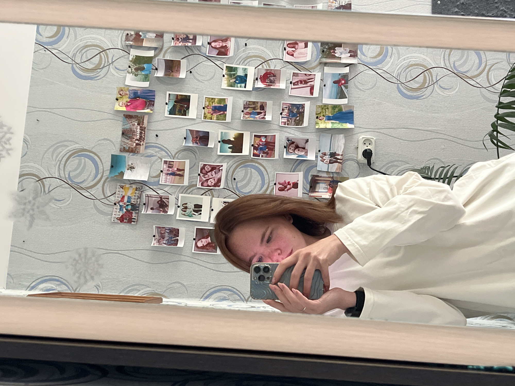 A person is taking a selfie in a mirror in Chaikovsky, Russia. The person is holding a smartphone and is wearing a white long-sleeved shirt. The background shows a wall with decorative wallpaper featuring a pattern of swirls and lines. Several small photographs are pinned to the wall. An electrical outlet is visible on the wall. A part of a potted plant is visible on the right side of the image. The room appears to be indoors.