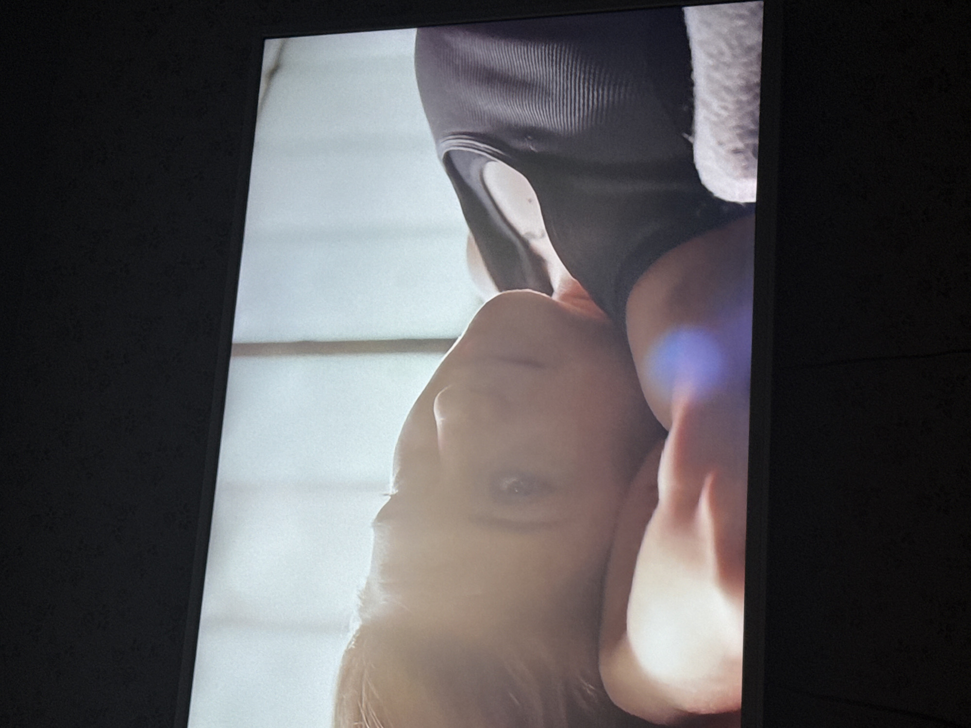 A close-up of a television screen shows a woman upside down, wearing a black tank top. The background is out of focus and appears to be a brightly lit room. The photograph appears to be taken in a dark room. There is no indication of this photograph being in Mytishchi, Russia.