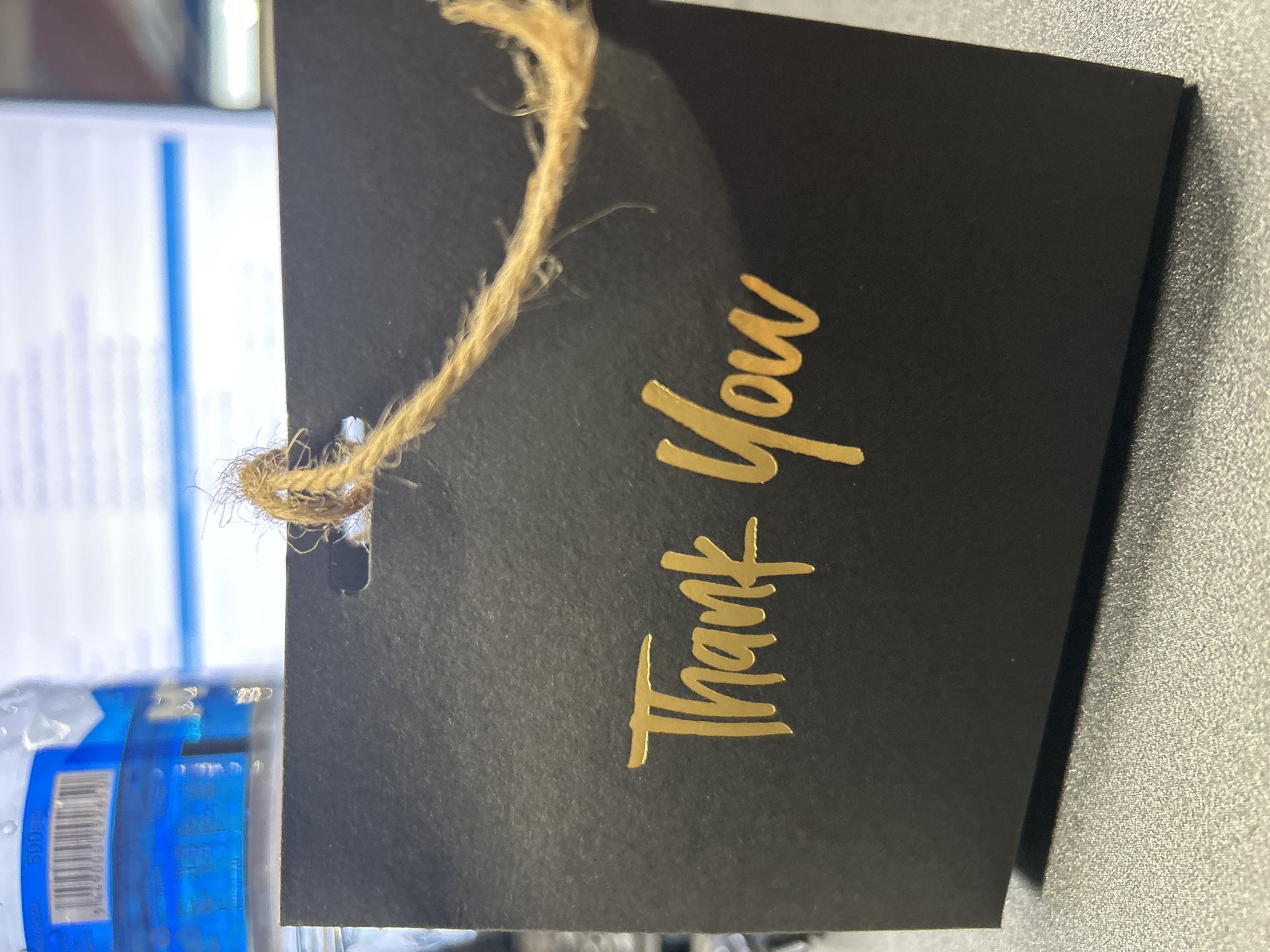 A black card with "Thank You" printed in gold foil is placed on a gray textured surface. A piece of twine is threaded through a hole at the top of the card. In the background, out of focus, is a blue and white label on a clear plastic bottle, and beyond that, a blurry office environment with computer monitors. The image is taken from a close-up perspective. The image does not provide any information about the location in the city of Tbilisi, Georgia.