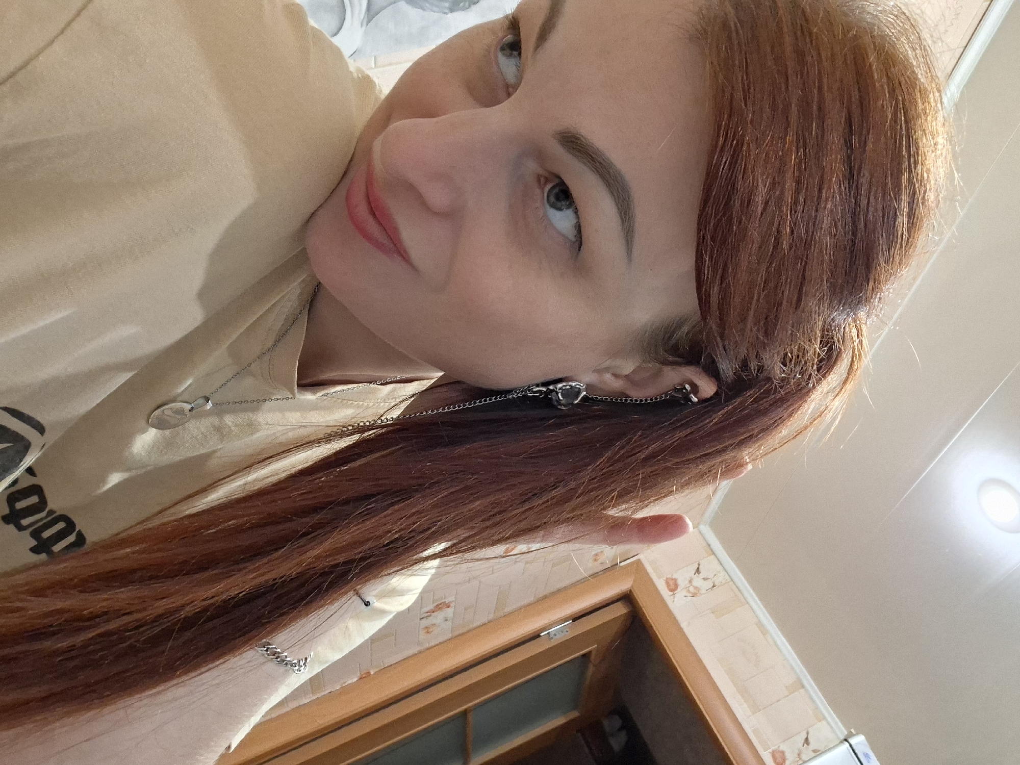A woman with long auburn hair pulled back into a ponytail is shown in a medium shot. She is wearing a tan t-shirt and several pieces of silver jewelry, including a chain necklace with a pendant, a chain bracelet, and an ear cuff with a dangling chain. Her makeup includes winged eyeliner and pink lipstick. She is looking towards the left side of the frame with a slight smile. In the background, there is a wall with floral wallpaper and a wooden doorway or cabinet. A white ceiling with a light fixture is visible at the top of the frame. The scene appears to be indoors, possibly in a home.