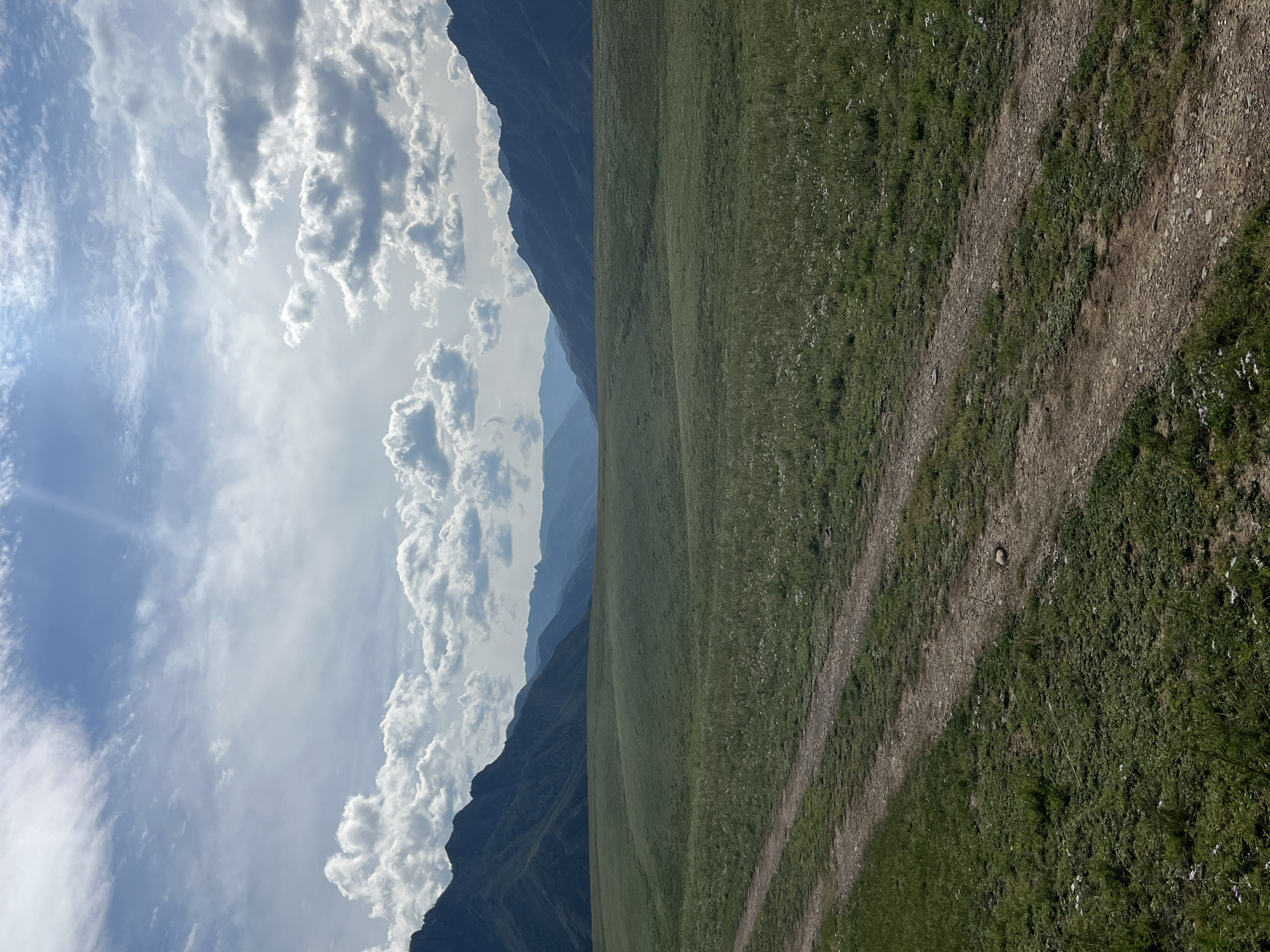 The image shows a wide, green, rolling meadow with a dirt track leading away from the viewer towards distant, hazy mountains. The sky is blue with scattered white clouds, and sunlight streams through in patches. The mountains in the background are layered, with the closest ones appearing darker and more defined, while those further away fade into a bluish haze. The foreground shows a well-trodden dirt path cutting diagonally across the grassy field. The overall impression is of a vast, natural landscape. The location is Malyi Yaloman, Russia.