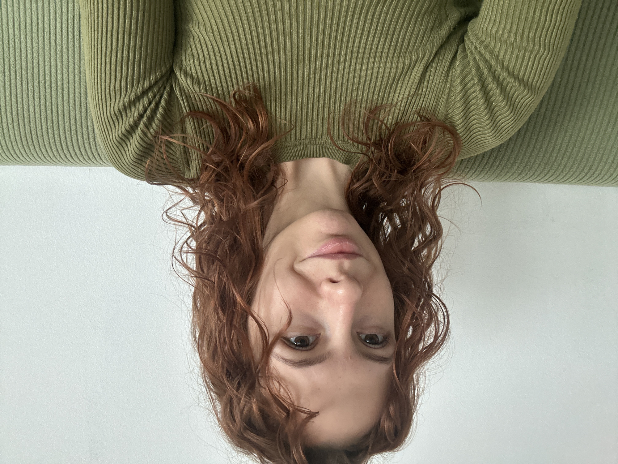 A selfie of a woman with reddish-brown, curly hair. She is wearing a light green ribbed sweater. The background is a plain white wall. The image is taken indoors. The location within the city of Novi Sad, Serbia cannot be determined from the visual information provided.