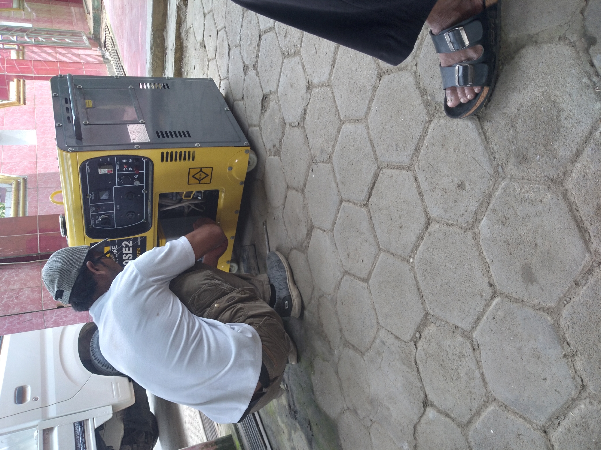 An adult male, wearing a white t-shirt, brown cargo pants, a grey baseball cap, and black slip-on shoes, is crouching down next to a yellow and grey portable generator. The generator has a control panel with various switches and a voltmeter. The man appears to be working on the generator. In the background, a white truck is partially visible. The ground is paved with hexagonal concrete blocks. The scene appears to be outdoors, possibly in a residential or workshop area, located in Pandak, Indonesia.