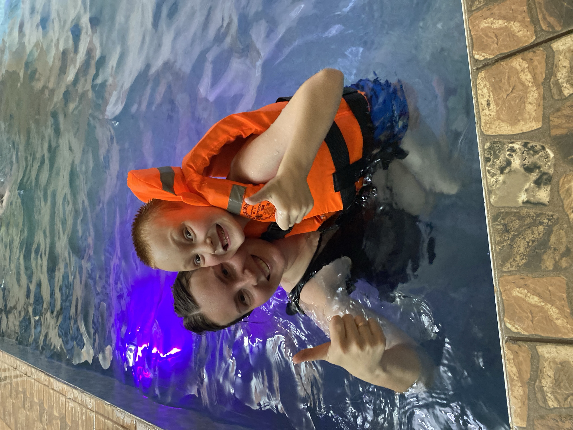 A photograph shows a woman and a young boy in a pool. The boy, wearing an orange life jacket, is holding onto the woman. Both are smiling and giving a "thumbs-up" gesture. The pool is surrounded by tiled edges. The location is Chaikovsky, Russia. The pool water appears to be illuminated with a blue light.