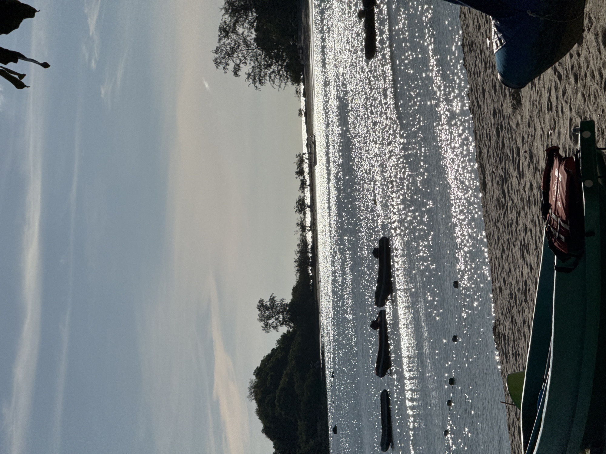 The image captures a sunny day at the beach, with a body of water reflecting the sunlight and creating a shimmering effect. In the foreground, a person's legs and feet, clad in blue shoes, are visible, resting on the sandy shore. Beside them, a life vest is attached to a green structure, possibly part of a boat or pier. The water is calm and dotted with what appear to be buoys or markers stretching out into the distance. The shoreline is lined with trees, suggesting a natural, somewhat secluded setting. The sky is bright blue with wispy clouds, indicating clear weather. The sun appears to be high in the sky, casting strong highlights on the water and illuminating the scene. The overall mood is peaceful and relaxed, typical of a day spent outdoors by the water.