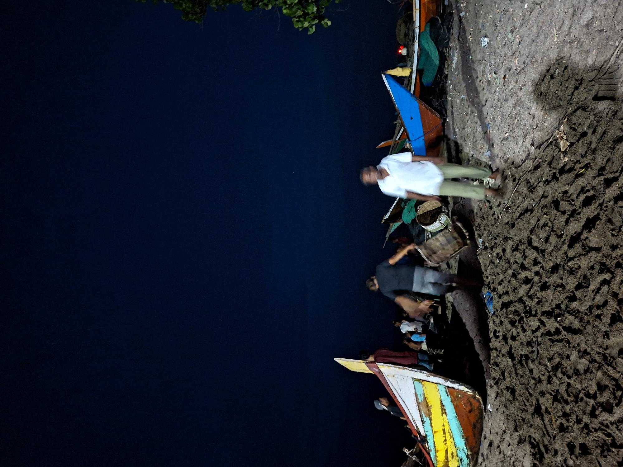 This is a low-light, long-exposure photograph taken at night on a beach in Lhokseumawe, Indonesia. Several fishing boats, painted in various bright colors, are pulled up onto the sandy shore. The sky above is a deep, dark blue, with a faint glow indicating ambient light. In the foreground, the dark, wet sand is textured with small waves and footprints.
There are several individuals present, some clearly visible and others blurred due to the long exposure. On the left, a boat with a colorful hull (yellow, red, blue, and white) is prominent. A person in a red shirt stands near its stern. Further back, other figures are visible, some appearing to be engaged in activities related to the boats.
In the center of the frame, a person in a dark shirt and grey shorts is blurred, carrying a woven cylindrical basket. To the right, a person in a white polo shirt and light green pants is also blurred, walking towards the right side of the image. In the background, more boats and figures can be seen, with a dark, leafy tree branch visible in the upper right corner. The overall scene suggests a moment of activity on the beach, possibly related to fishing operations after dark.