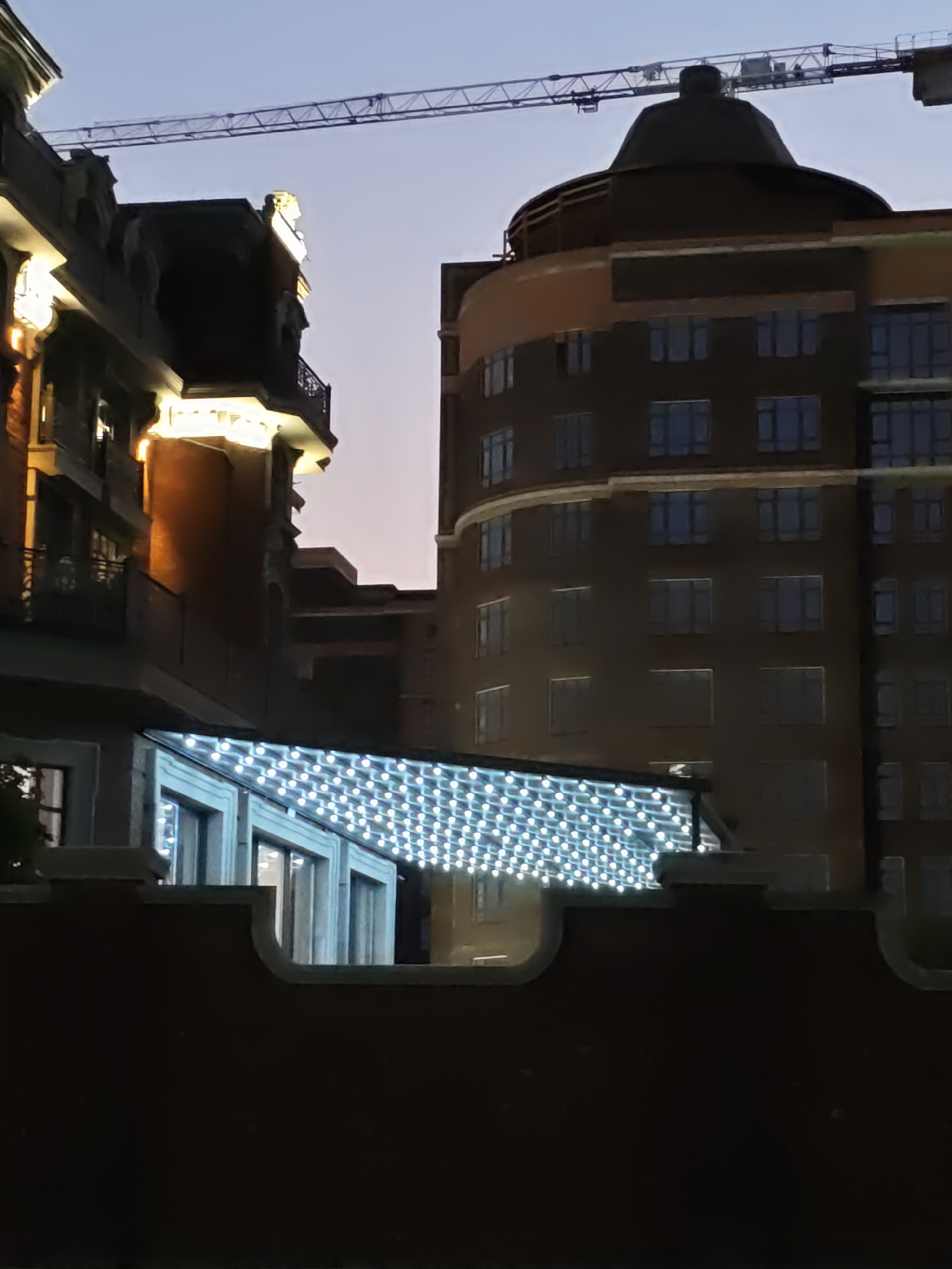 The photograph captures an outdoor scene at dusk or night, featuring an ornate brick building on the left and a modern, curved building on the right. A construction crane is visible in the background, extending across the sky. The building on the left has detailed architectural features, including decorative molding and illuminated windows. A covered walkway with numerous string lights runs along the lower portion of the scene, connecting to the building on the left and possibly extending towards the building on the right. The lower foreground is dominated by a dark, textured wall with a curved top edge. The overall impression is a blend of historical and contemporary architecture with ongoing construction. This scene is located in Rostov-on-Don, Russia, specifically at the Rostov Arena stadium development.