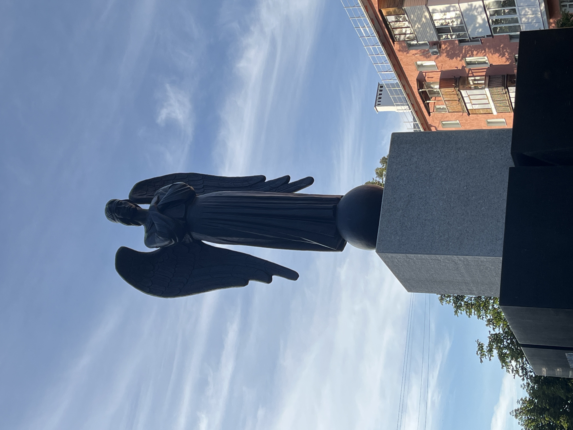 A dark statue of an angel with wings is positioned on top of a gray rectangular pedestal. The angel is looking down with its hands clasped in front of its chest. The statue is located in Nizhny Novgorod, Russia, in front of a coral-colored building with balconies. The sky is blue with wispy clouds.