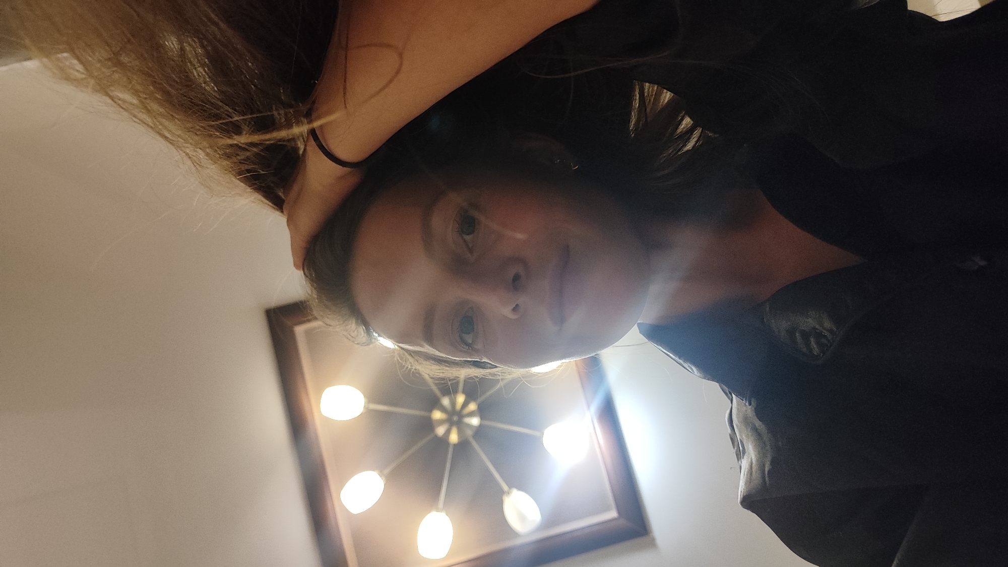 A photograph taken from a low angle shows a person with long, light brown hair, their left arm raised with a black elastic band visible on their wrist. The person is wearing a dark colored garment, likely a shirt or jacket. The person's face is visible with light eyes. In the background, a framed light fixture with multiple bulbs is visible against a light-colored wall. The scene's location in Chaikovsky, Russia, cannot be determined from the image.