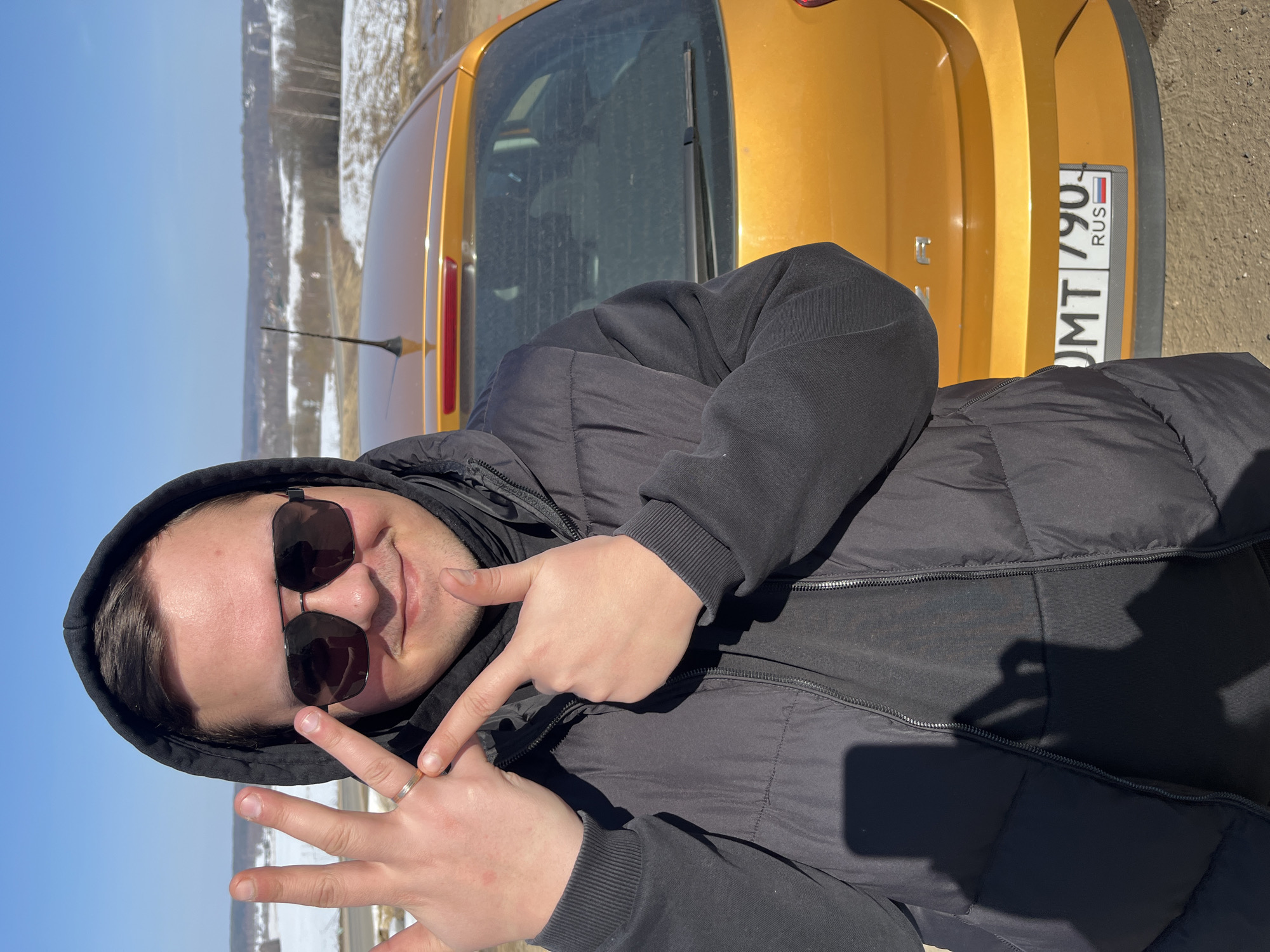The image displays a male subject standing outdoors in Zubovo, Russia. The subject, a young adult male with dark hair partially visible under a black hood, wears a black hooded puffer jacket over a dark garment and dark sunglasses. He faces the camera with a slight smile. His left hand is raised with fingers splayed, revealing a thin gold-colored ring on the ring finger. His right hand is raised with the index finger pointing towards his left hand. Directly behind the subject is the rear of a bright orange hatchback vehicle, showing its rear window, a single wiper, and taillights. A black antenna is visible on the vehicle's roof. The license plate, partially visible, reads "UMT 790." with "RUS" and the Russian flag emblem. The immediate ground is exposed earth or gravel. The background features a clear blue sky above a wide, undulating landscape with distant hills, partially covered in snow, and scattered leafless trees. A road is discernible in the mid-distance. The scene is illuminated by strong natural light.