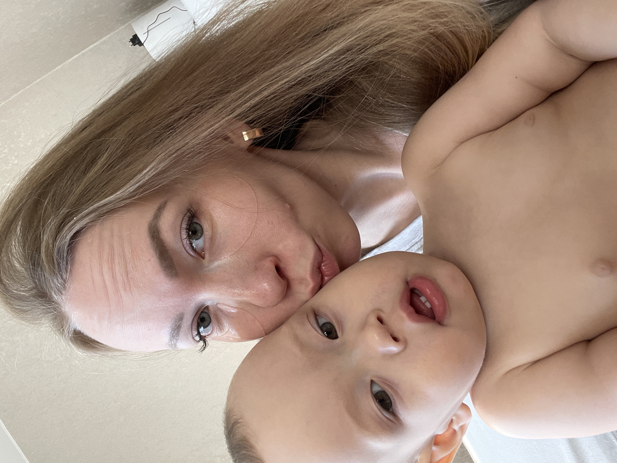 A light-skinned woman with blonde hair and a gold earring poses with a light-skinned baby, likely in an indoor setting, based on the light and lack of exterior elements. The location is potentially Perm, Russia. The woman is close to the baby, possibly kissing its head or cheek. The baby has its mouth slightly open and is looking towards the camera. The background is neutral, appearing to be a wall or a plain surface. There is a white object near the top left corner, possibly a fixture on the wall.