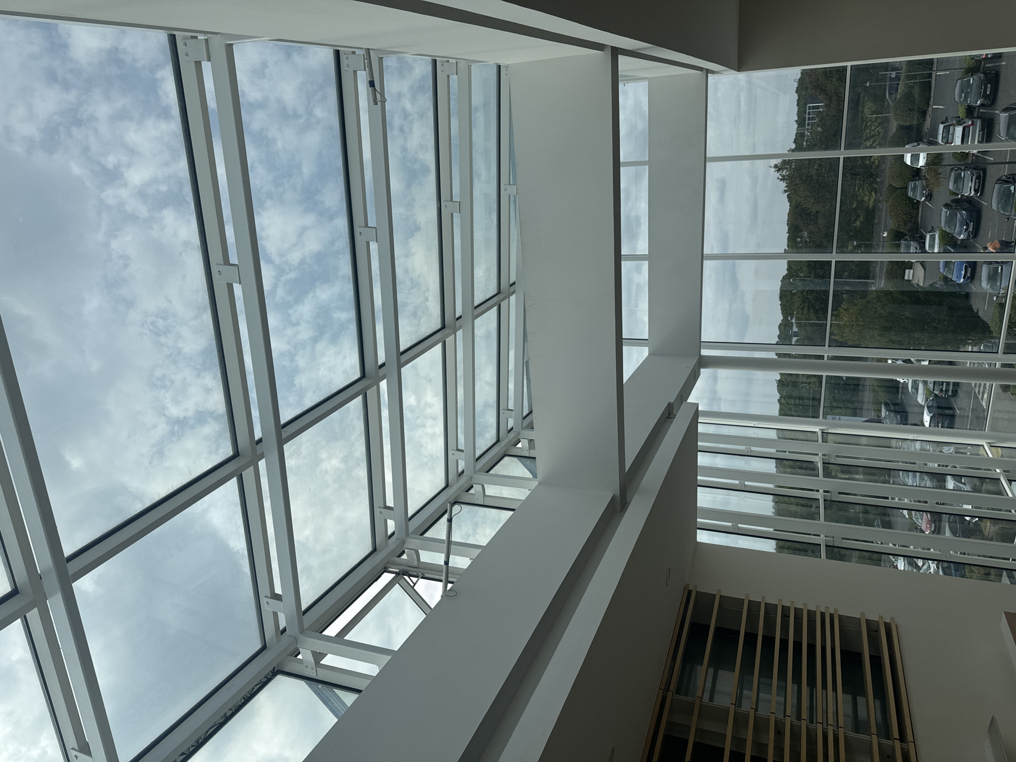 The image shows a view looking up at a modern glass and white-framed atrium ceiling with a cloudy sky visible through it. Below the atrium, large windows look out onto a parking lot filled with cars and a wooded landscape under an overcast sky. There are no visible people in the frame. The building appears to be an office or commercial space. The location is identified as Unknown, Ireland.