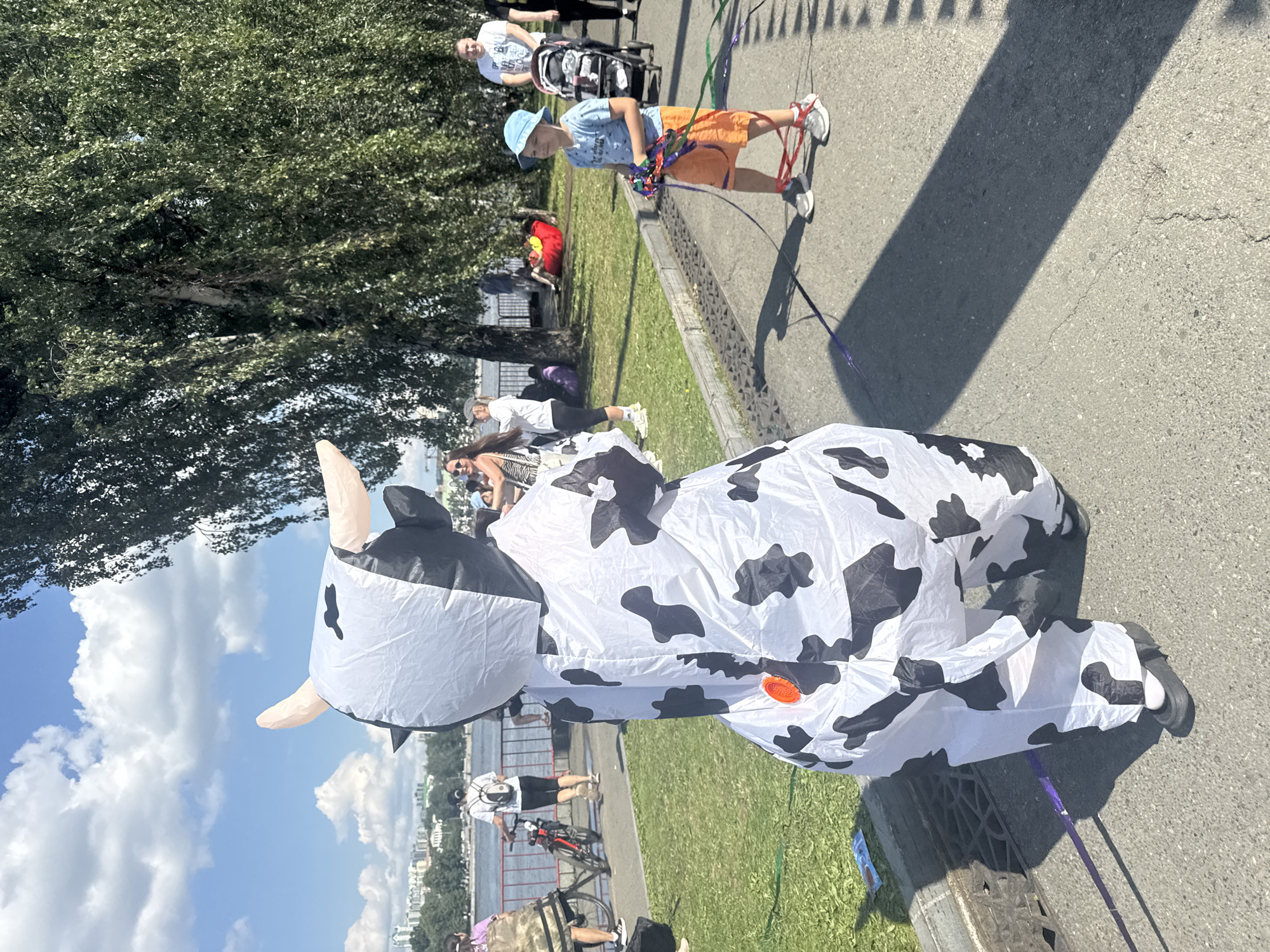 An inflated cow costume stands in the foreground of a sunny outdoor scene, facing away from the camera. The costume is white with black cow spots and has large horns. Behind the cow, a young boy in a blue hat and orange shorts is holding a string with colorful streamers. In the middle ground, several people are walking and sitting on the grass. A woman in a zebra-print top and a man pushing a stroller are visible. In the background, there is a body of water with buildings on the far side, and a line of trees along the right side of the image. The location is the embankment of the Iset River in Yekaterinburg, Russia, likely near the Boris Yeltsin Presidential Center, given the urban landscape and prominent trees.