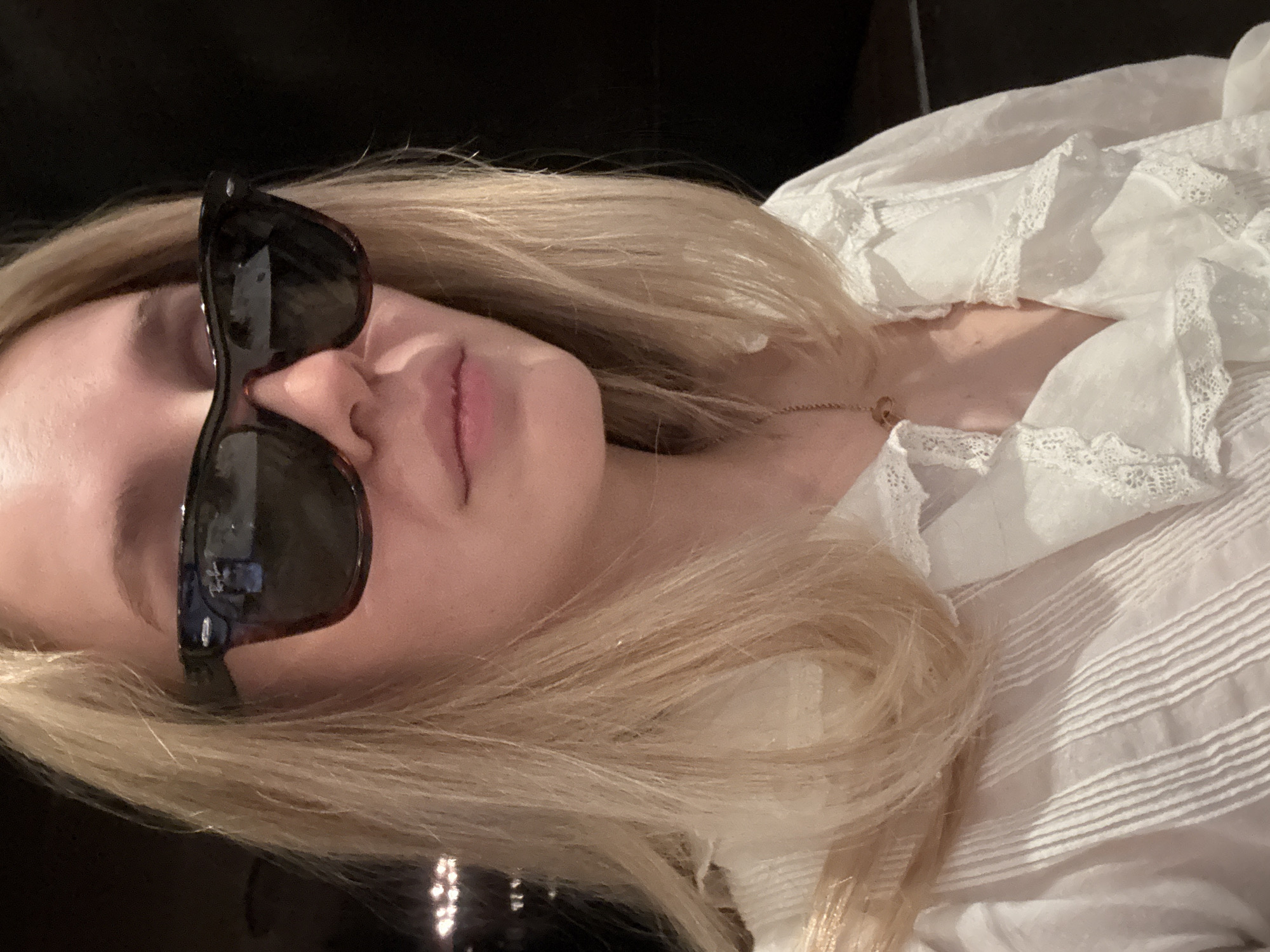 A pale-skinned woman lies on a dark surface wearing a white ruffled blouse and sunglasses. She has long, blonde hair. A gold necklace is visible on her neck. The lighting is dim, and the background is dark, obscuring any environmental details that could place her in Mexico City.