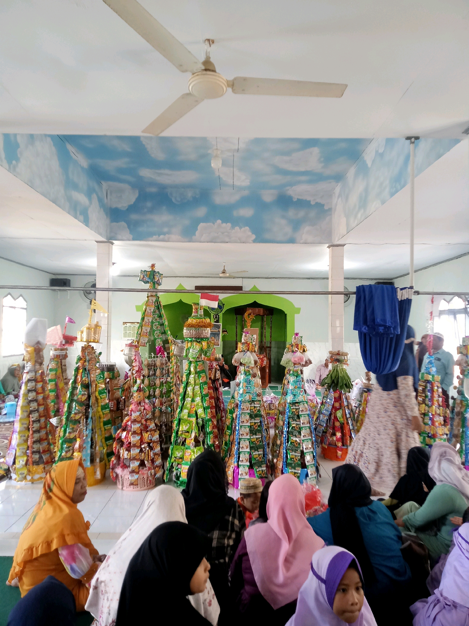In a building with a white ceiling adorned with a painted sky mural and a ceiling fan, a religious gathering takes place. The room is illuminated by natural light filtering through windows and overhead lights. The floor is tiled with white squares. The focal point of the scene is a collection of tall, cone-shaped structures decorated with various packaged goods, resembling offerings or decorations. These structures are arranged in rows.
Seated on the floor in the foreground are several individuals, predominantly women wearing headscarves in various colors including orange, black, white, and pink. They are facing towards the decorated structures. Some are looking down, while others are looking forward. To the right, standing individuals are also visible, some in the background. A blue cloth is draped on a horizontal bar in the middle ground. A green archway is visible in the background, and above it, an Indonesian flag is displayed. The overall atmosphere suggests a communal event or ceremony.