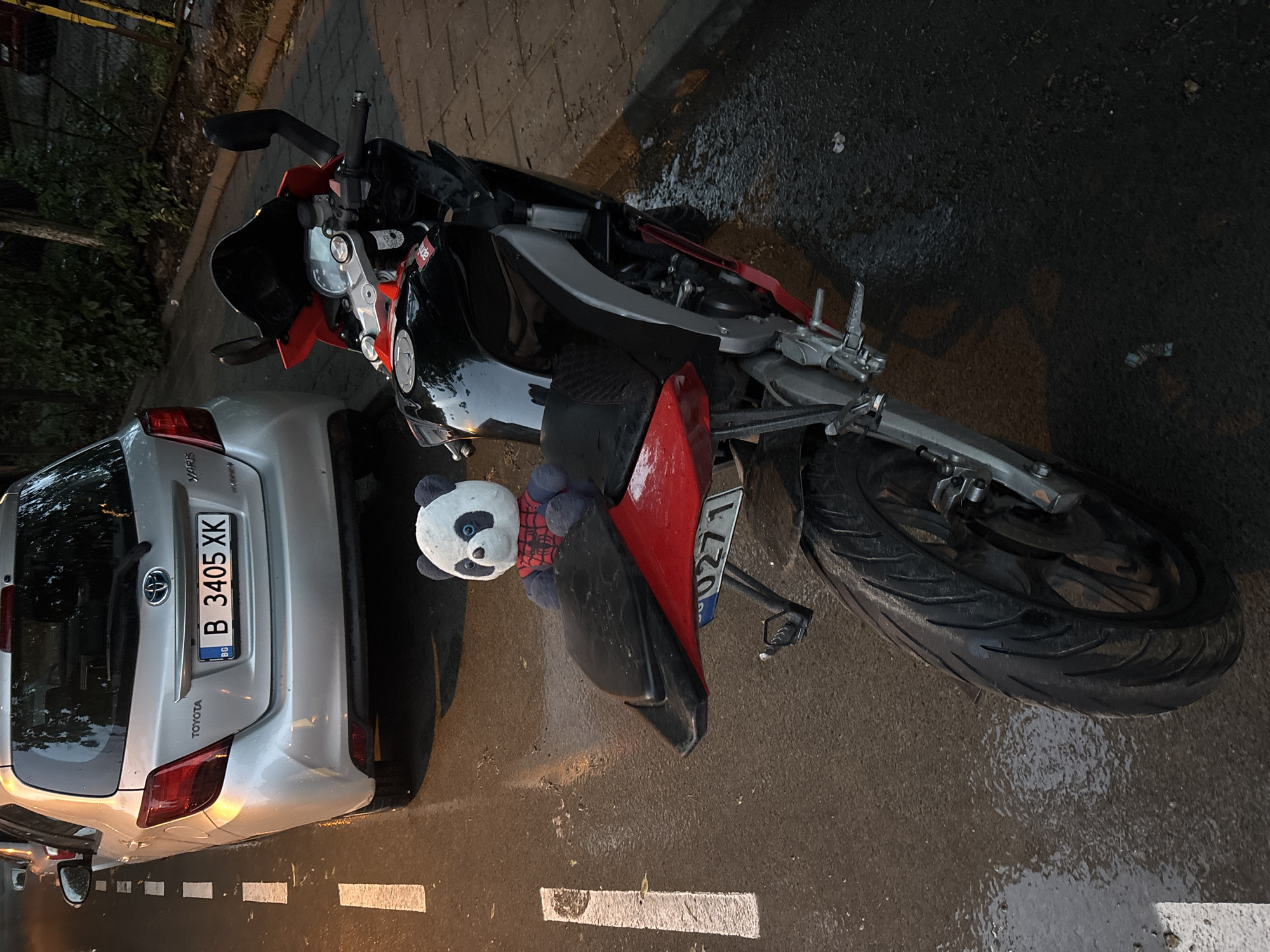 The image captures an overhead view of a red and black Aprilia sports motorcycle parked on a wet asphalt street, next to a silver Toyota Yaris hatchback. The ground is visibly wet with puddles, indicating recent rain. The scene appears to be at dusk or night, illuminated by artificial light.
The motorcycle is positioned at an angle, with its front end near the rear of the car. A white and black panda plush toy, wearing a red Spiderman shirt, is placed on the motorcycle's rear seat, leaning against the back fender.
Visible text on the car includes "TOYOTA" and "YARIS" on the tailgate, and the Bulgarian license plate "B 3405 XK" with the "BG" country code. This confirms the location context of Bulgaria, with "B" indicating Sofia, not Varna as stated in the prompt, which is an important distinction. The motorcycle also has a license plate, partially obscured by the plush toy, showing "0271" at the bottom. An "Aprilia" sticker is visible on the motorcycle's body. White dashed lines are painted on the wet asphalt road. The motorcycle's rear tire shows wet tread.