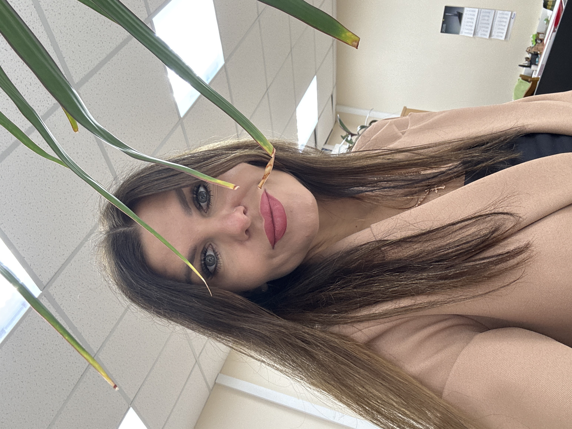 A woman with long brown hair, fair skin, and light-colored eyes, wearing a tan blazer and a black top, looks directly at the camera. Green plant leaves are prominent in the foreground, partially obscuring her face. The background includes a white, tiled ceiling with recessed lighting, and a wall with a calendar and other office items. The image appears to be taken indoors, potentially in an office setting. There is no evidence to verify the location is Chaikovsky, Russia.
