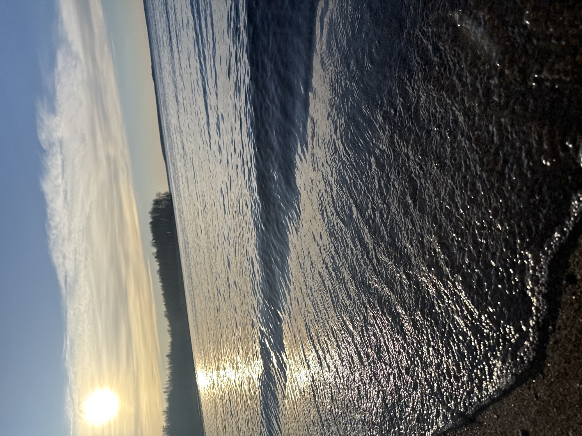 The image shows a sunset over a body of water, likely a lake, with the sun low on the horizon. The shoreline is visible on the right edge of the frame, where the water meets the sand. The water's surface is rippled with waves, reflecting the sunlight. In the distance, a dark line of trees and land are visible across the water. The sky is a mix of blue and white clouds, with a golden hue from the setting sun.
It is not possible to identify the exact location in the city of Glubokoe, Russia based on this image alone.