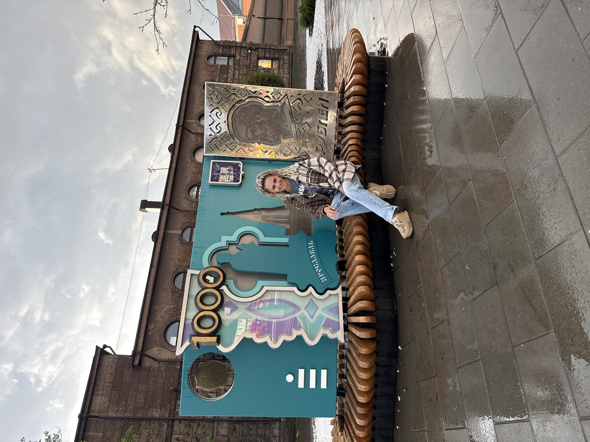 A medium shot depicts a blond woman sitting on an ornate bench in Заволжье, Russia. The woman wears a flannel shirt, blue jeans, and tan shoes. She is positioned in front of an upright, rectangular structure composed of a blue panel with decorative cut-outs and inscriptions in Russian, topped with a metallic panel featuring an embossed portrait. The bench seating is constructed from curved, stacked wooden slats. The ground consists of wet, grey paving stones. In the background, a weathered, multi-story brick building is visible, with circular windows on its upper levels. Above, the sky is overcast with heavy clouds.