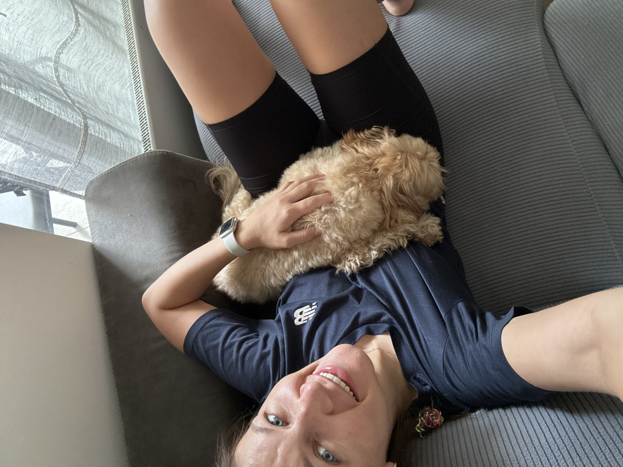 A selfie taken from a low angle looking up at a Caucasian woman lying on a grey couch in a brightly lit room, holding a fluffy, light brown dog on her chest. The woman is smiling broadly with blue eyes, wearing a dark blue t-shirt with a white logo and black cycling shorts. She has a white smartwatch on her left wrist and is holding the dog with her left hand. The dog is resting comfortably on her. Behind the woman is a window with sheer, patterned curtains, suggesting an interior setting. The couch is light grey with a horizontal lined pattern. The location is unknown beyond the possible city.