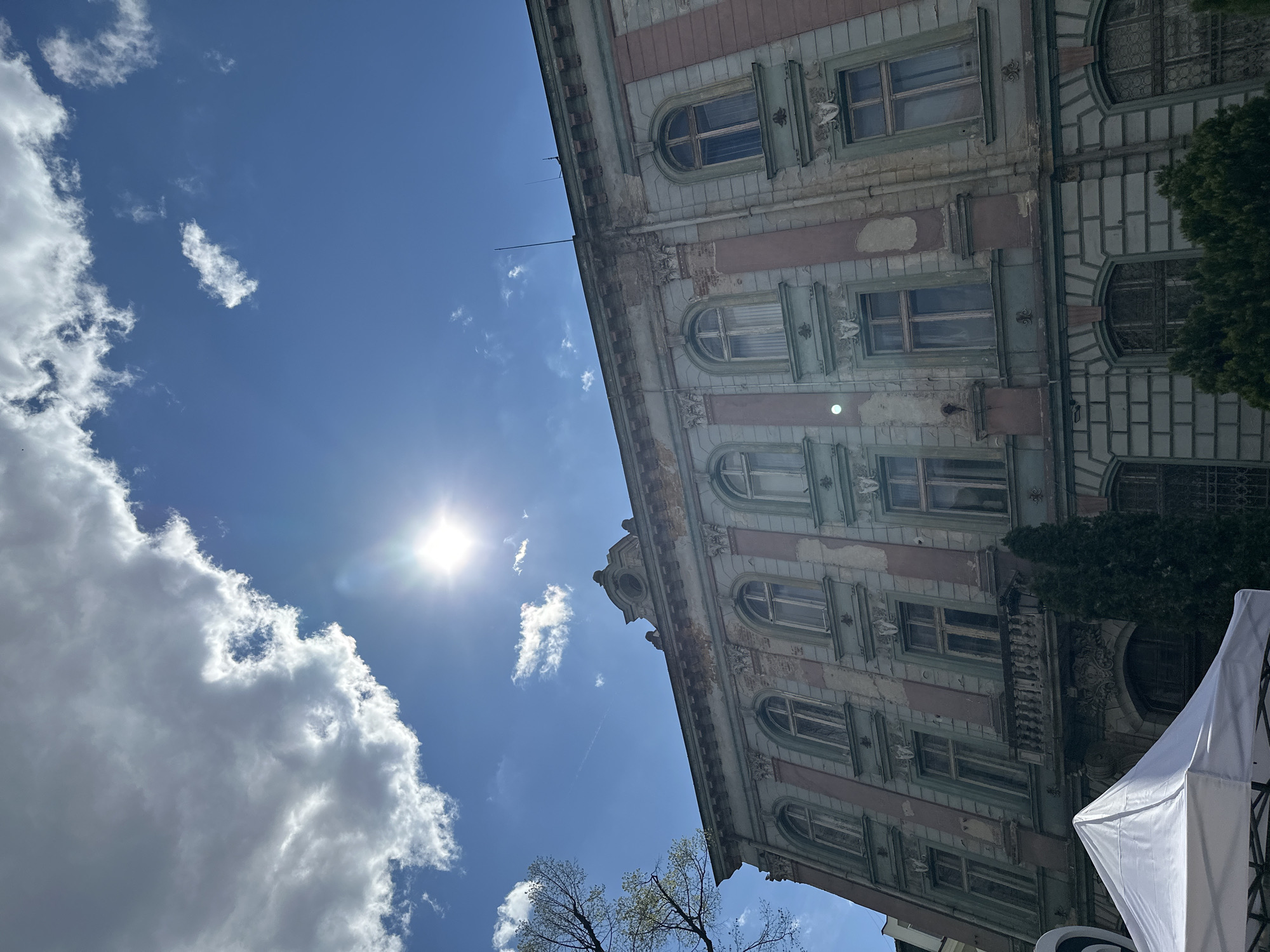 The image depicts a tilted view of an ornate building in Plovdiv, Bulgaria, under a bright blue sky with scattered clouds and the sun shining. The building features multiple stories with arched windows, decorative moldings, and a faded color scheme of pink, green, and gray. The building's facade shows signs of age and disrepair, including peeling paint and weathered surfaces. The sky is mostly clear, with some puffy white clouds and what appears to be a contrail from a jet plane. Part of a white tent or awning is visible on the lower right of the image. A tree with budding leaves is visible in the lower-middle foreground and some larger greenery is visible attached to the building on the right.