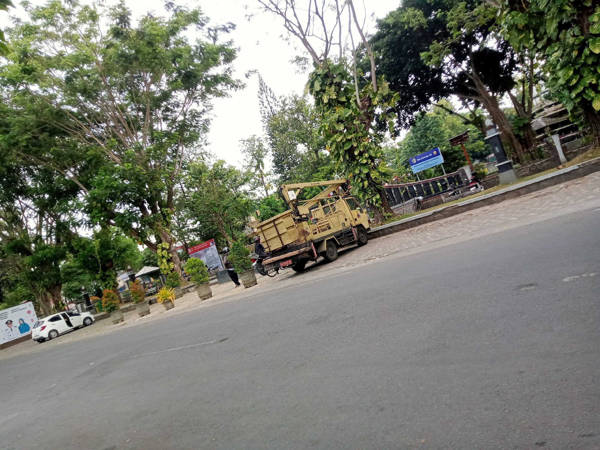 The image depicts a street scene in Tulungagung, Indonesia, with a focus on a yellow truck equipped with a crane. The truck is parked on the side of a road, which appears to be asphalt and in moderate condition. To the left of the truck, a white car is parked, and further left, a large banner displaying portraits of two individuals is visible.
The truck's crane is extended upwards, suggesting it might be in operation or prepared for use. The truck itself is weathered, with its bed appearing to be a container. Behind the truck and the white car, there is a sidewalk lined with planters containing small trees and shrubs. Beyond the sidewalk, lush green trees and vegetation form a dense backdrop, with some buildings and a fence visible through the foliage.
A few people are present in the scene. One individual, wearing a dark helmet and jacket, is seated on a motorcycle parked behind the truck. Another person, dressed in dark clothing, is standing near the planters to the left of the truck. A blue sign with text in Indonesian is mounted on a post behind the fence, indicating a restricted area or information. The sky is overcast, suggesting a cloudy day. The overall environment suggests an urban or semi-urban setting with significant greenery.
