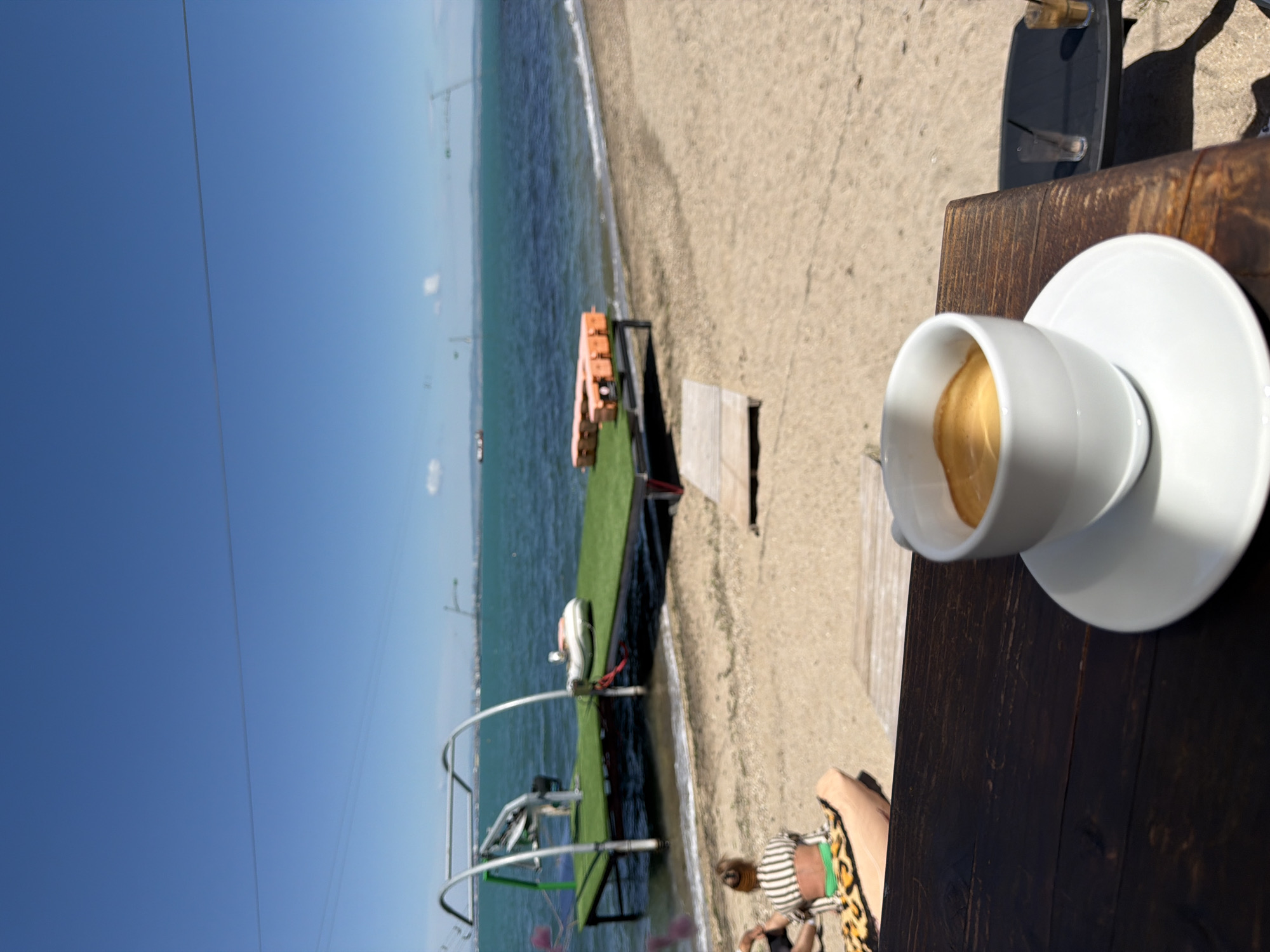The image depicts a cup of coffee on a dark wooden table next to a beach. In the background, there is a sandy beach and blue water with a dock. A person's legs and part of their clothing are visible near the table. The scene appears to be set in a cafe or restaurant located near the beach in Burgas, Bulgaria.