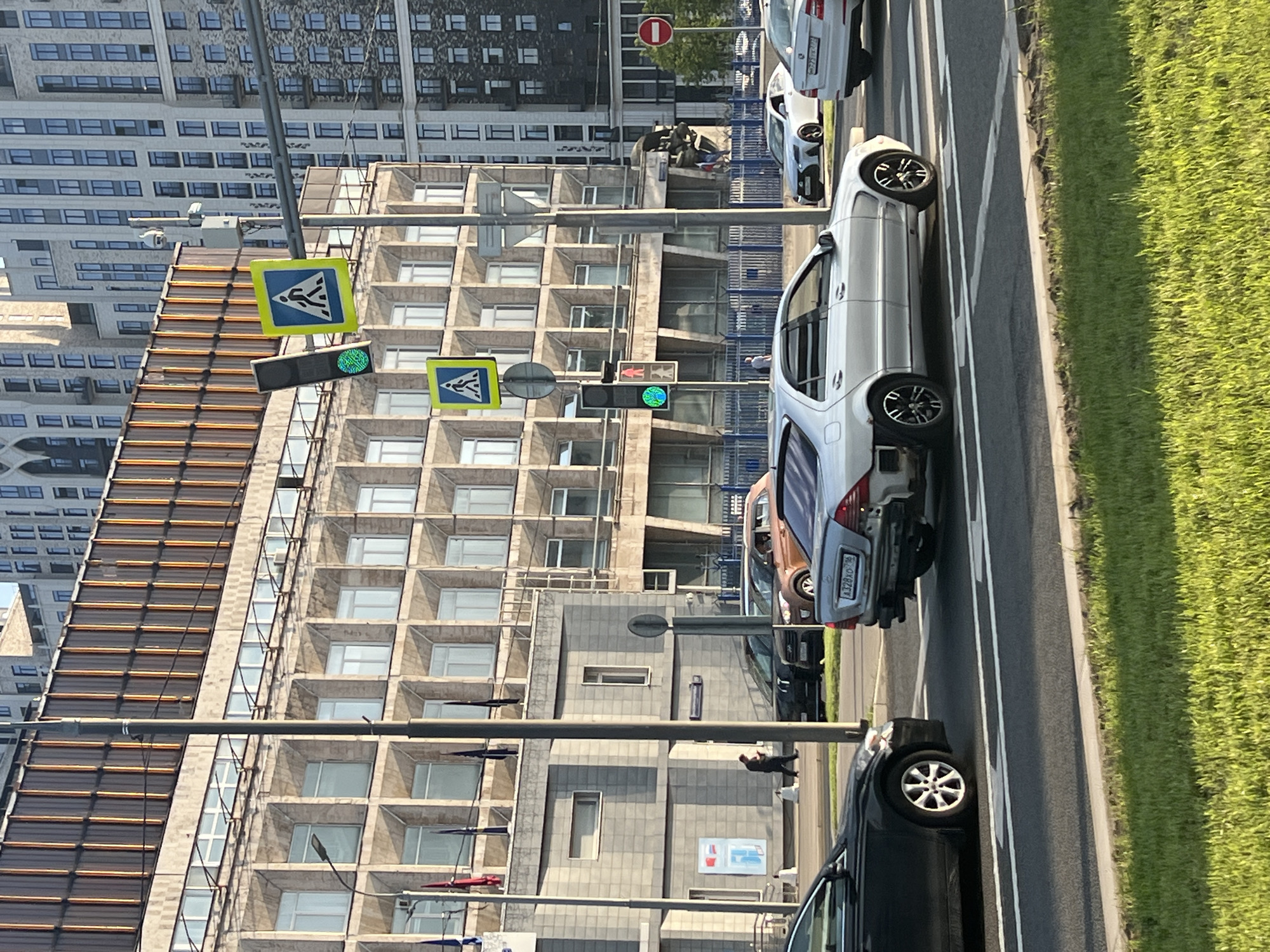 The image shows a street scene in Moscow, Russia, featuring a multi-lane road with several parked cars along the right side. A patch of green grass borders the road. Pedestrian crosswalk signs are visible on lampposts, with the accompanying traffic lights showing green. Buildings of varying styles are in the background, including a modern structure with a grid-like facade and a taller skyscraper in the distance. A few individuals are discernible on the sidewalk.