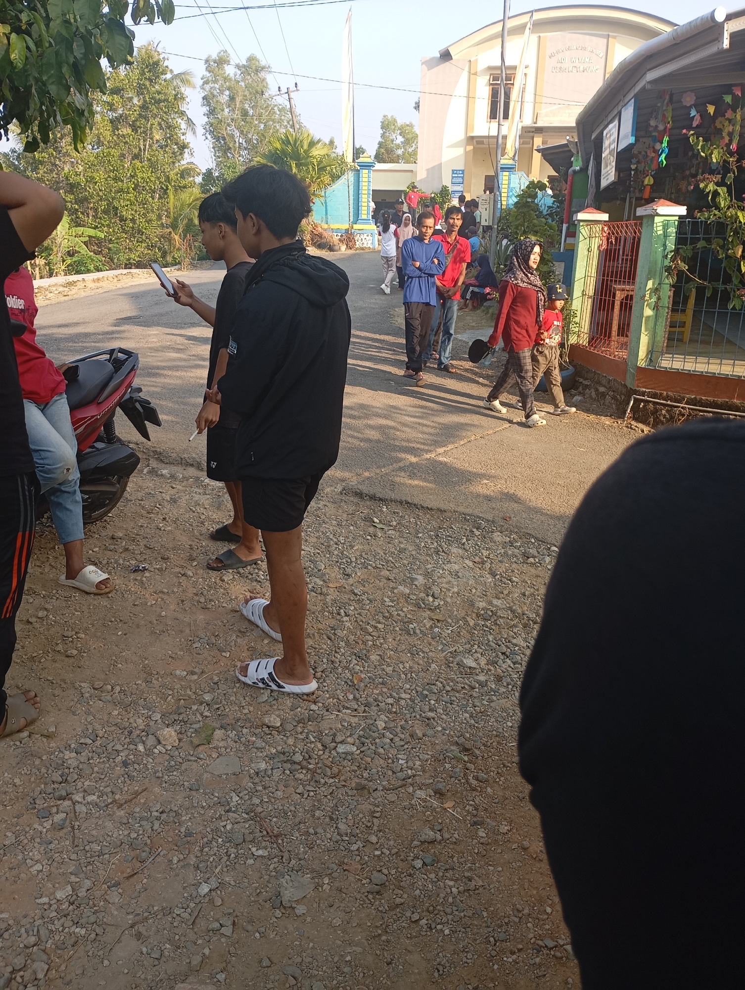 The image depicts a street scene in Ketepung, Indonesia, with several individuals present. In the foreground, on the left, a person in a red shirt and blue jeans is partially visible, standing next to a red motorcycle. To their right, two young men are standing, one of whom is holding a smartphone. One of these men is wearing a black jacket and shorts, while the other wears a black t-shirt and shorts. Another young man is seen to the right, wearing a black jacket. Further down the street, a group of people are walking. This group includes a woman in a headscarf and maroon top, a child in a red shirt, and other individuals dressed casually. A building with a curved roof, possibly a public institution, is visible in the background, with signage in Indonesian. The environment appears to be a semi-rural or suburban area, with trees and vegetation lining the street. The ground is a mix of paved road and gravelly dirt. The lighting suggests it is daytime, with the sun casting shadows.