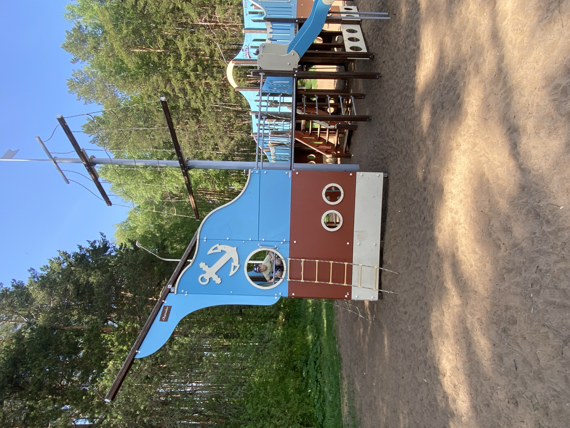 The image shows a playground structure in the shape of a pirate ship located in Chaikovsky, Russia. The structure features blue and brown panels, an anchor design, mast, rigging, porthole windows, a ladder, and a slide. A child is visible inside one of the portholes. The playground is located near a wooded area, with tall trees visible in the background. The ground around the playground is covered in sand. The sky is blue.