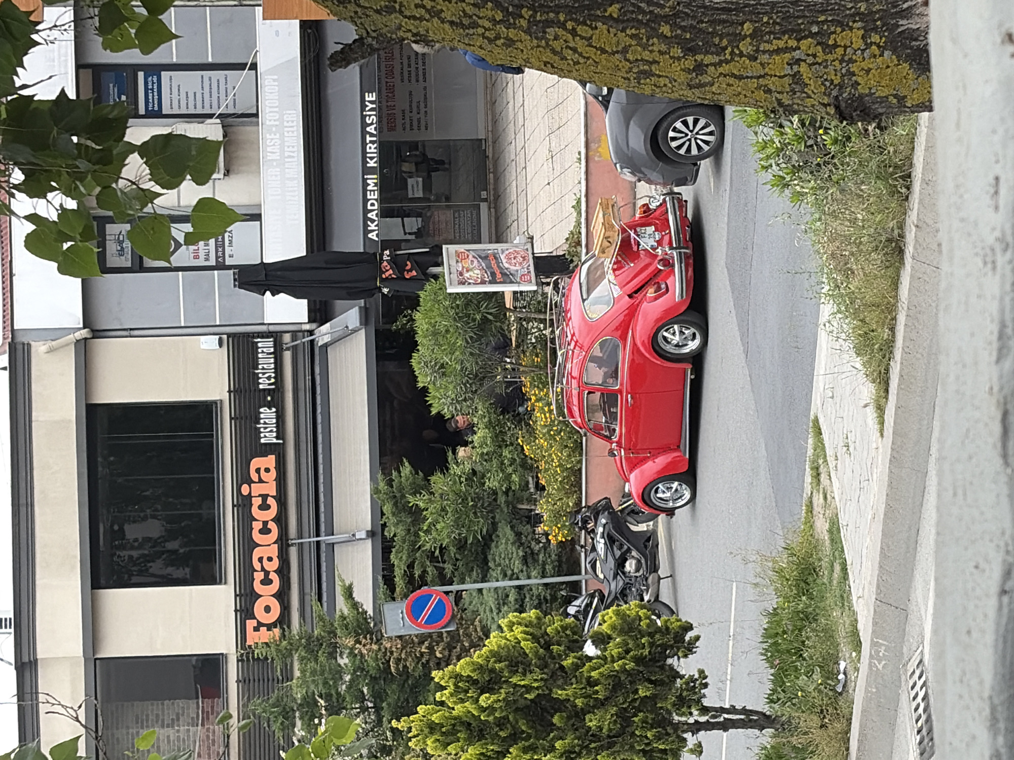 The image shows a street in Esenler, Turkey. On the left is a building with a restaurant called "Focaccia pastane restaurant" and a business providing "TİCARET SICIL DANIŞMANLIĞI" services. Next to it is "AKADEMİ KIRTASİYE" selling stationery. A red vintage car is parked on the street, next to a parked black motorcycle. There's a blue and red parking sign nearby. Pedestrians are visible in the background. A tree trunk and branches partially obscure the view on the right and left of the image.