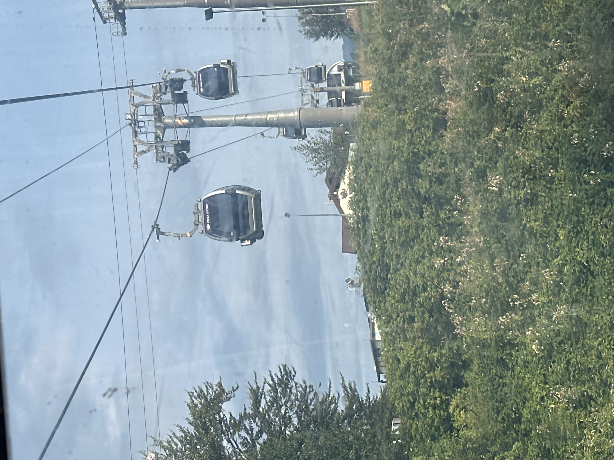The image shows a ski lift or cable car system with multiple cabins suspended from overhead cables. The cabins are dark, enclosed units with windows, and some have the text "ROSA KHUTOR" visible on them. The cable cars are supported by large metal towers. In the background, there are trees and some buildings, including a building with a pitched roof and what appears to be a brown and white logo. The sky is overcast with patches of blue. The scene is likely located in Estosadok, Russia, given the visible text on the cable cars. The overall impression is of a scenic mountain or resort area.