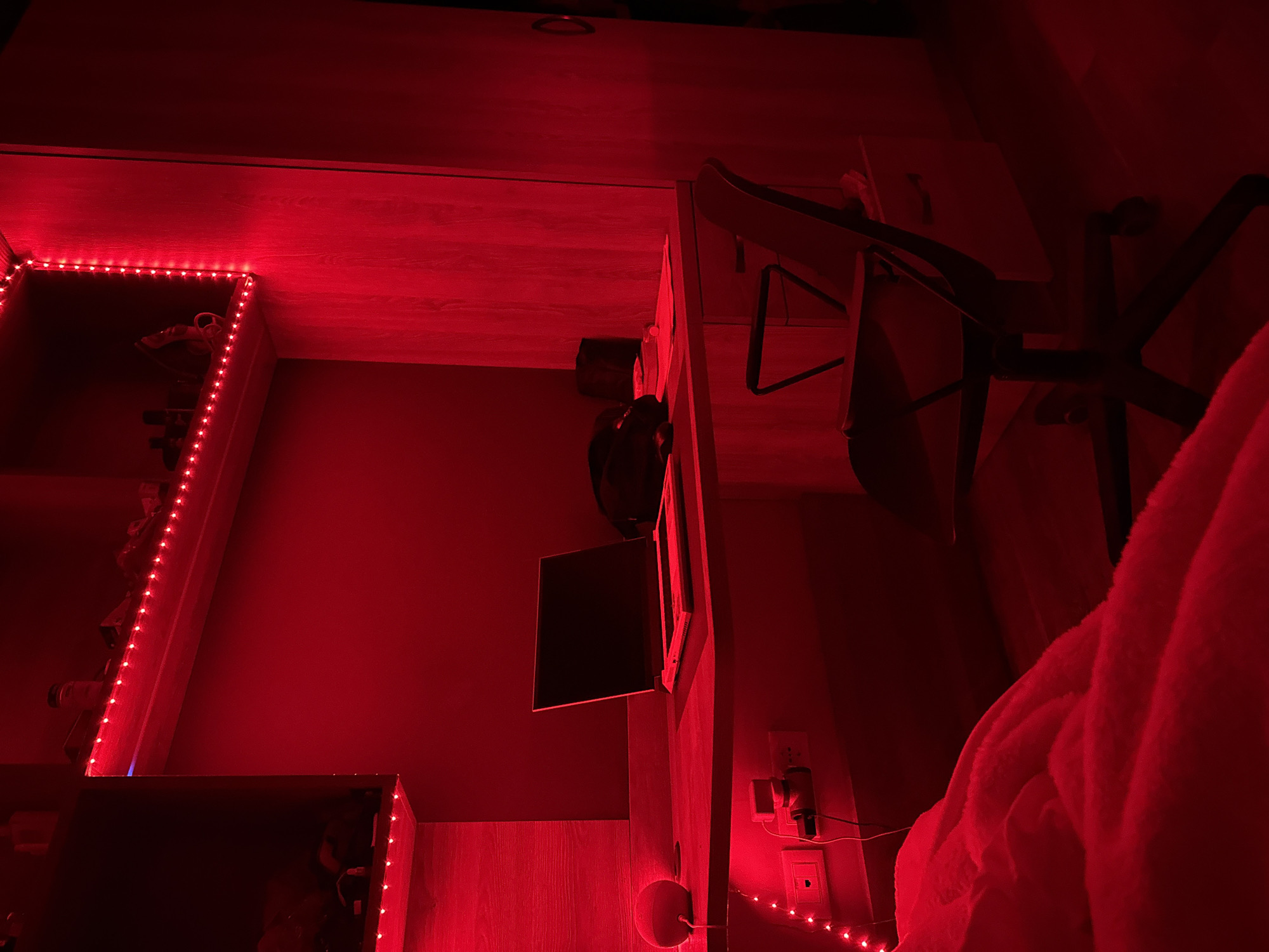 A room is illuminated by red LED strip lights, with an overhead shelf unit running along one wall. A desk with a chair is visible in the right side of the frame. A television or monitor is mounted on the wall. Electrical outlets and a Google Home device are plugged in near the right side. There is a light-colored fluffy blanket or robe is in the foreground. The specific location in Milan, Italy is not discernable from the image.