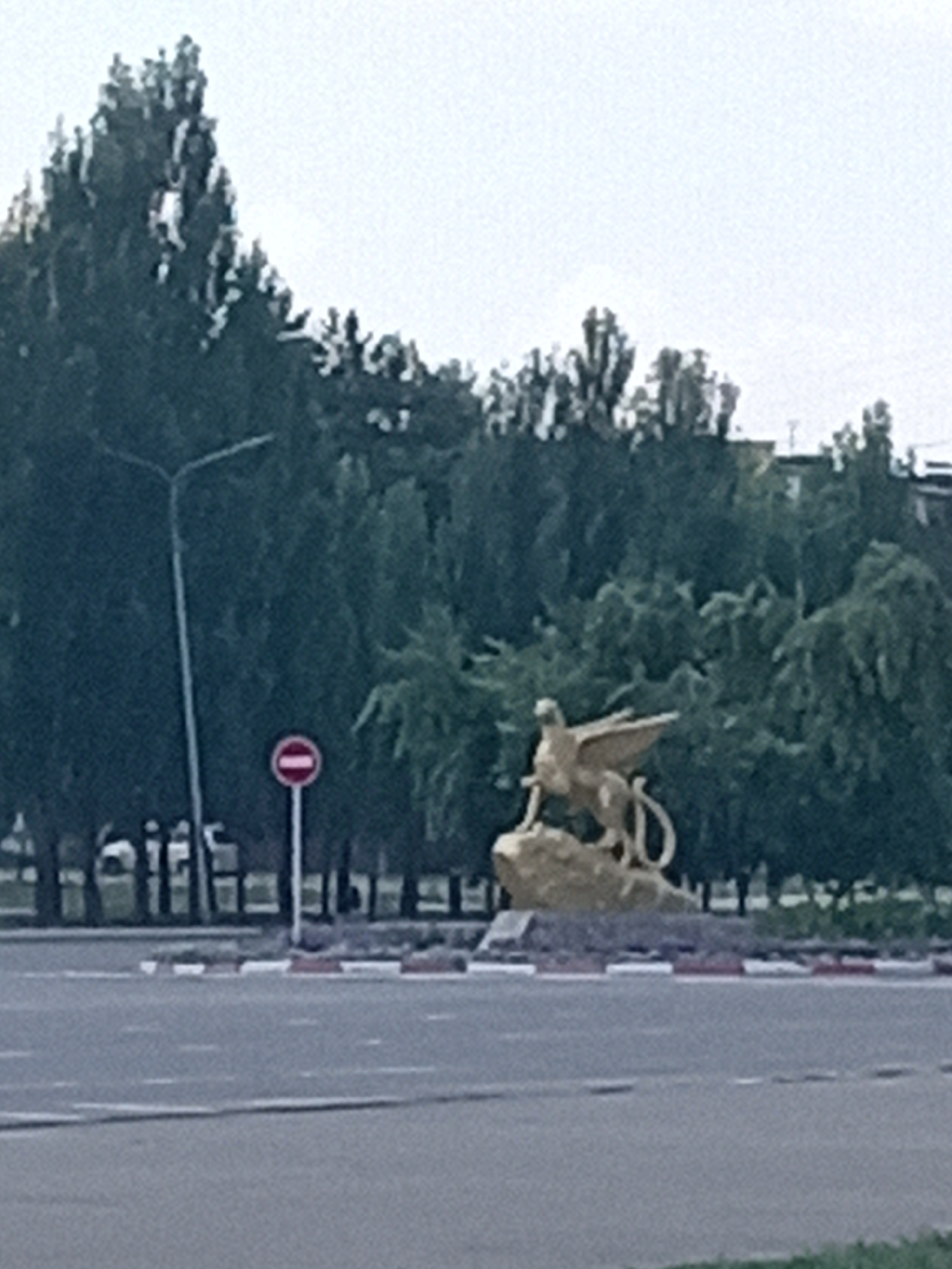 The image shows a golden statue of a winged creature, possibly a griffin, atop a rock in a city setting. The statue is positioned on a roundabout. Behind the statue, there are many trees and some buildings visible. A street lamp with a "no entry" sign is visible to the left of the statue. The foreground shows a road with white markings and a red and white striped curb. The overall scene appears to be a street in the city of Rudny, Kazakhstan, with a monument as the central focus.